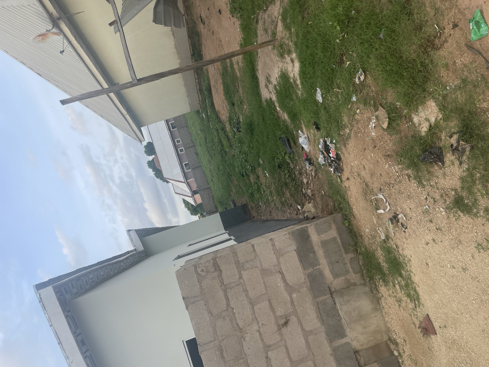 The image depicts an outdoor scene in the city of Jalingo, Nigeria. In the foreground, there is a partially constructed building with a concrete block foundation. The building has a light-colored exterior wall and a dark base. Adjacent to this building is another structure with a light yellow exterior and a corrugated metal roof. A wooden pole stands vertically in front of this second building. The ground is a mixture of dirt and patchy grass, with scattered debris including plastic bags, wrappers, and what appears to be discarded construction materials. In the background, more buildings with similar roofing styles are visible. The sky above is partly cloudy. The overall impression is that of an unfinished or developing area with some litter.