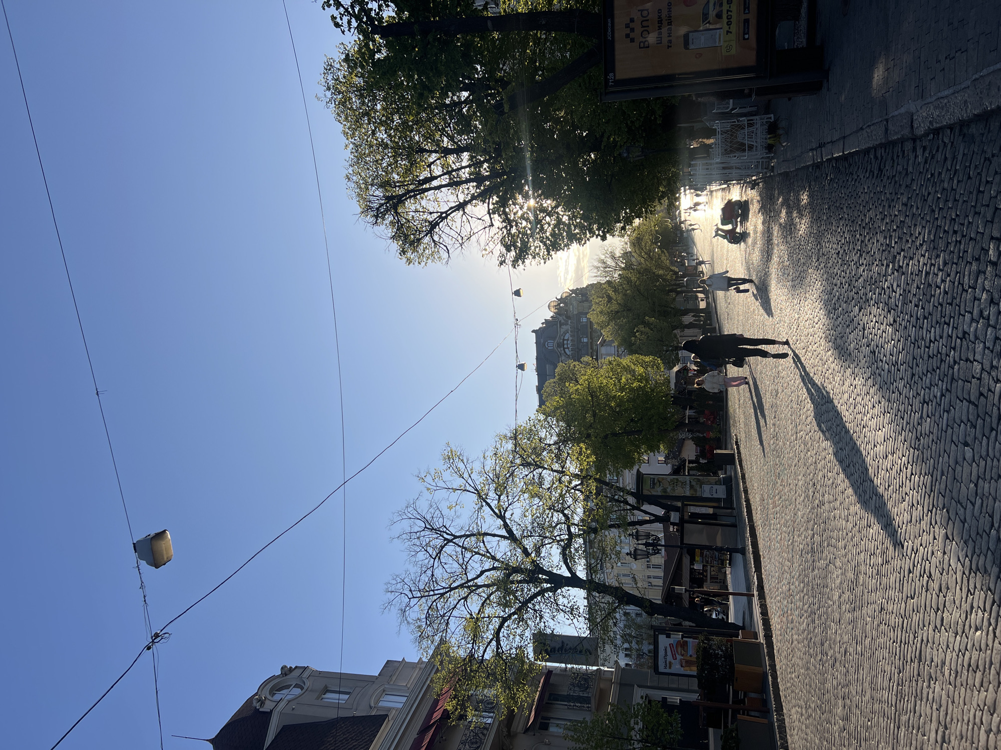 The image presents a sun-drenched urban street scene, likely in Odesa, Ukraine, during either early morning or late afternoon, as evidenced by the low angle of the sun, long shadows, and prominent lens flare. The sky is clear and blue.
The street itself is paved with distinct cobblestones, indicating a historic or pedestrian-friendly area. Numerous pedestrians are visible, mostly silhouetted against the bright light, walking along the path. Some outdoor café seating, with tables and stacked chairs, can be seen along the street, suggesting local businesses and leisure activities.
Mature trees with green foliage, some still showing sparse branches, line the street, casting dappled shadows. Traditional-style buildings with ornate windows and detailed architecture are visible on both sides. Overhead, a network of utility wires or tram lines spans the sky, with a suspended light fixture or electrical component.
Visible text includes an advertisement on a stand for "Bond," with Ukrainian text "Швидко та надійно" (Quickly and reliably) and a partial phone number "7-007-0...". Another sign further down reads "Навігатор" (Navigator), and part of a "Radisson" sign is discernible on a building. The scene depicts a lively city environment under bright, clear skies.