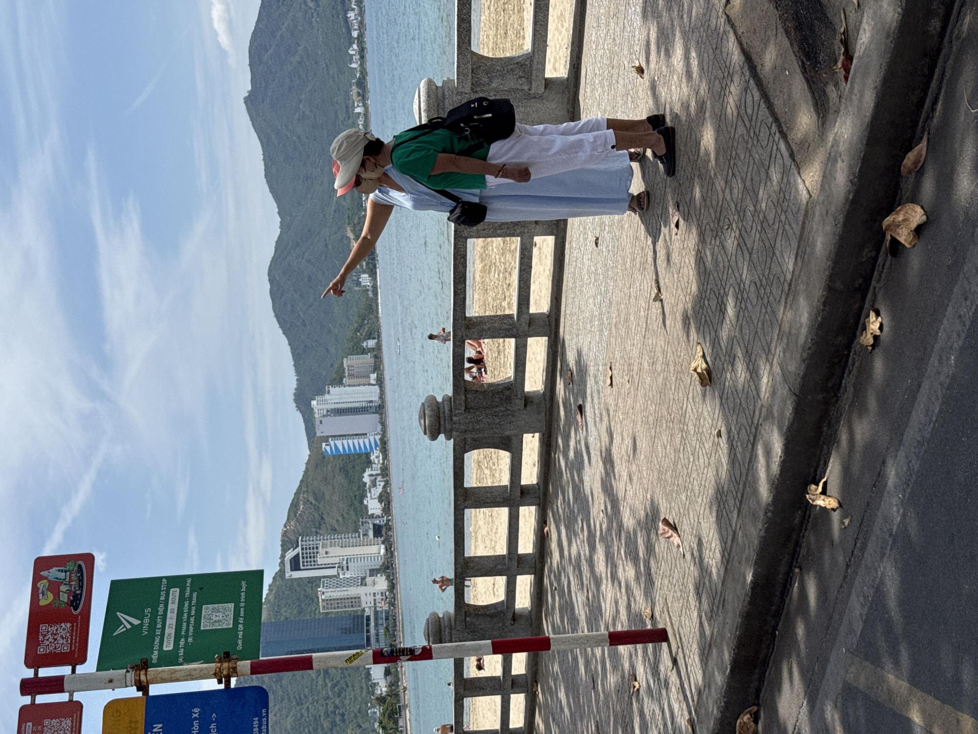 The image shows two women standing on a paved sidewalk overlooking a beach in Nha Trang, Vietnam. One woman, wearing a light blue dress and a red cap, is pointing towards the ocean. The other woman, in a green top and white pants, stands beside her. In the background, there is a sandy beach with a stone railing, the turquoise ocean, and a cityscape with tall buildings and green hills. A red and white striped pole with signs is on the left side of the frame. The scene is set on a sunny day.
