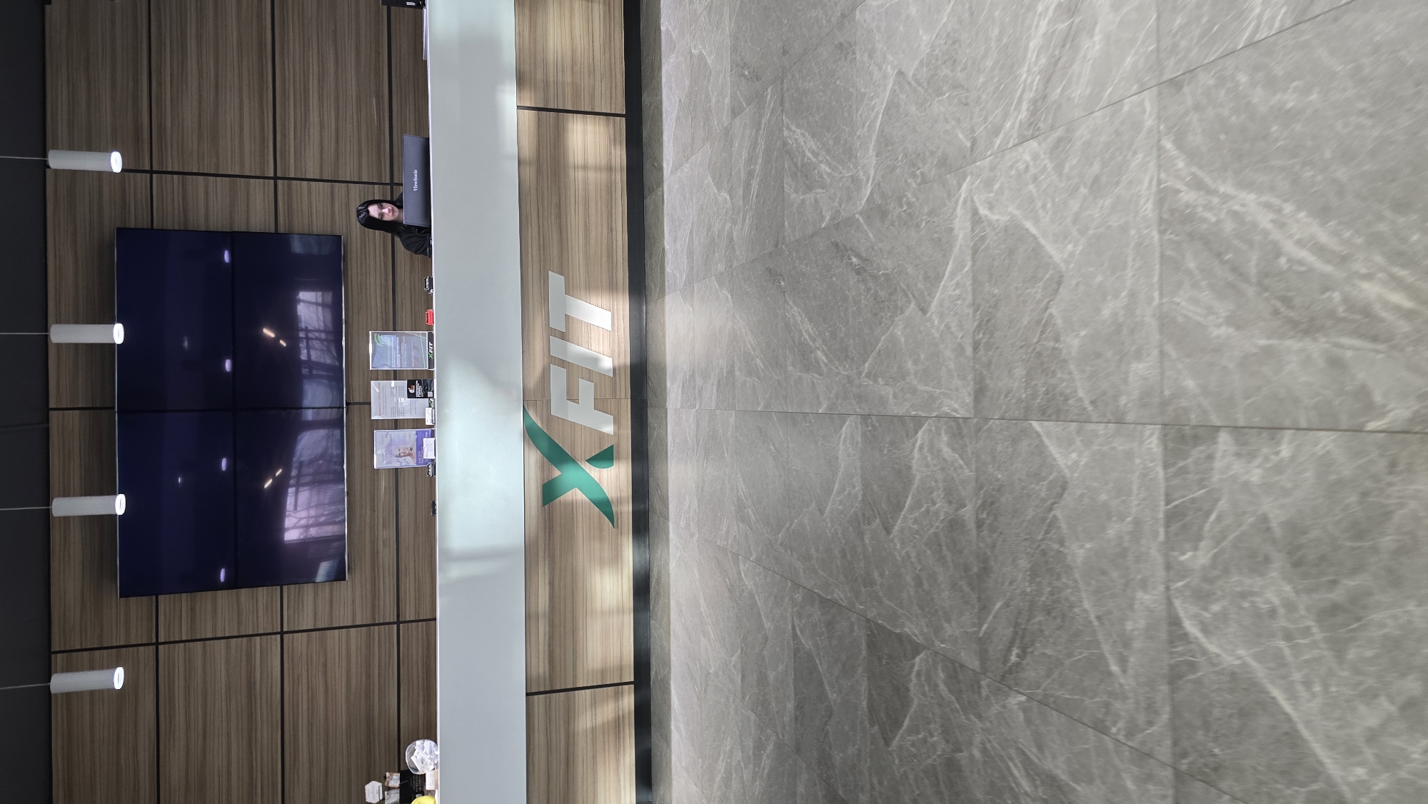 The image shows the interior of a reception area, possibly a gym or fitness center. A woman is visible behind a reception desk. The desk has the "X-FIT" logo on its side. Behind the desk are wall panels with screens displaying content. Above the desk are cylindrical light fixtures. The floor is covered with large gray marble-like tiles. The location is likely in Kaliningrad, Russia.