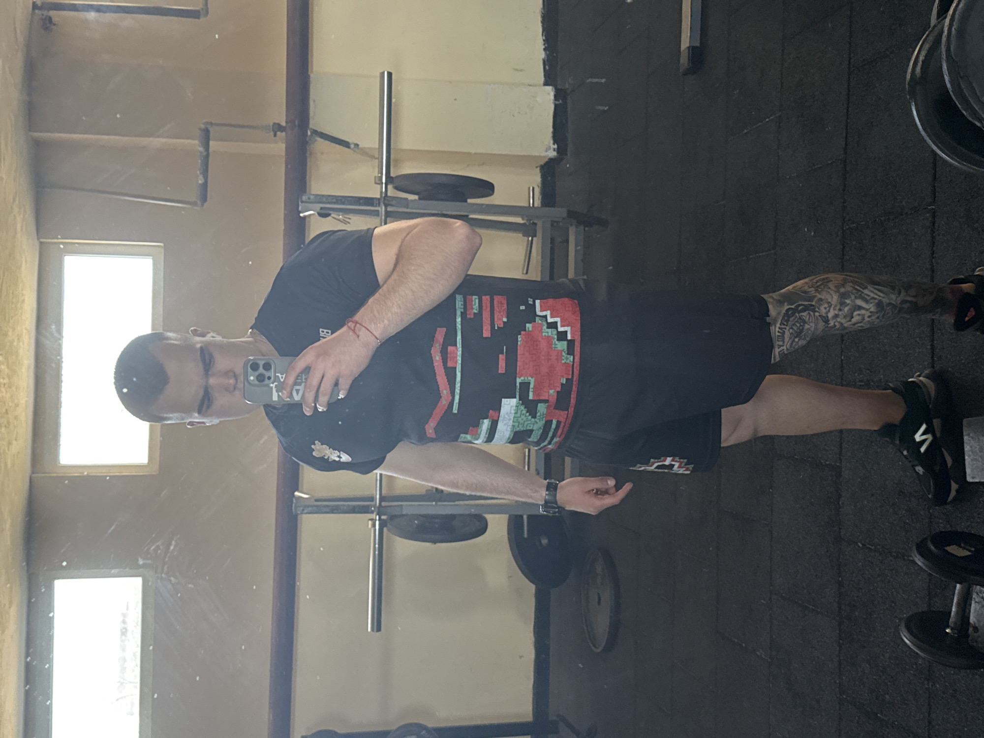 A man is taking a selfie in a gym. The location appears to be an interior space with gym equipment, including weight racks and dumbbells, visible in the background. The floor is covered with dark rubber mats. The man is wearing a t-shirt, shorts, and sneakers. He has tattoos on his leg. Natural light enters the gym through windows on the wall to the left. The location of the gym in the city of Unknown, Unknown is not provided.