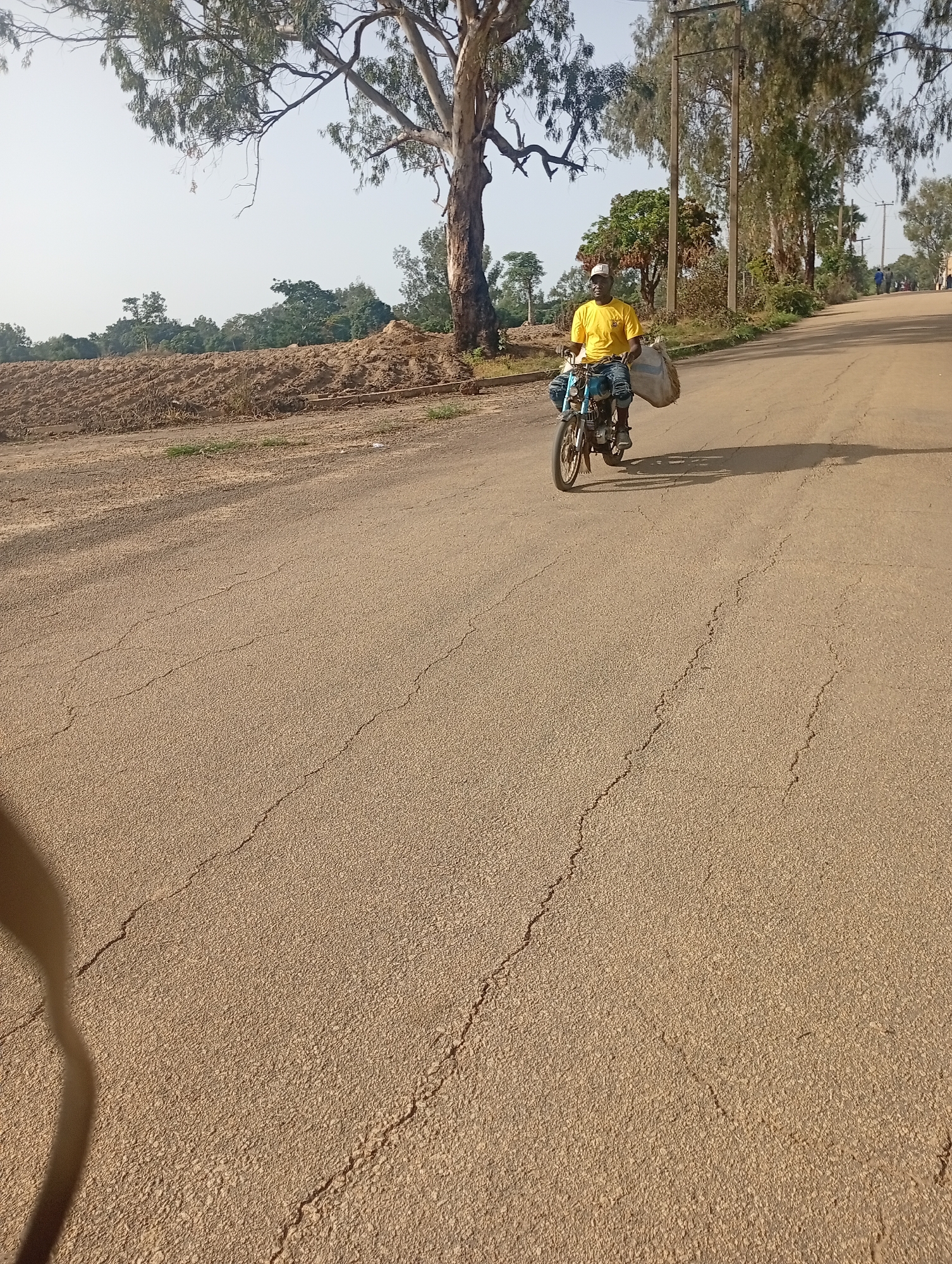 The image depicts a man riding a motorcycle on a paved road. The man is wearing a yellow t-shirt and blue jeans, and he is carrying a large bag on his back. The motorcycle appears to be a light model. The road is flanked by trees and vegetation. In the background, there are buildings and utility poles. The sky is clear and bright. This scene is likely in Jos, Nigeria, a city known for its temperate climate and hilly terrain. The road surface shows signs of wear and tear, with visible cracks. The overall atmosphere of the image is one of everyday life in an African city, with a focus on transportation and local scenery.