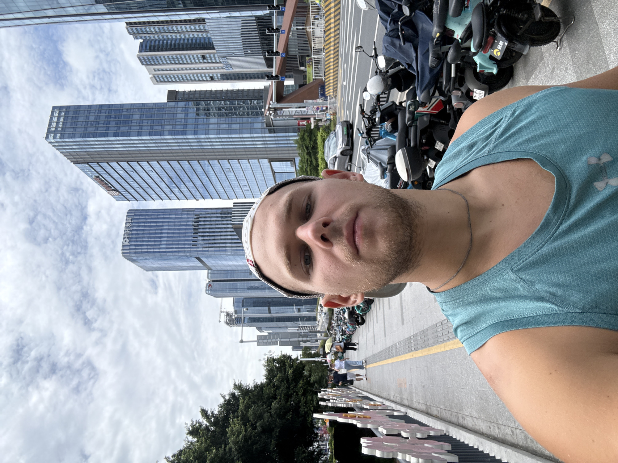 A man wearing a teal tank top and a baseball cap takes a selfie while standing on a sidewalk in Tianhe District, China. He is looking directly at the camera. In the background, several modern skyscrapers rise into a cloudy sky. A street is visible with parked motorcycles and a car. The sidewalk has decorative fencing and a yellow line marking its edge. A few pedestrians are walking in the distance.