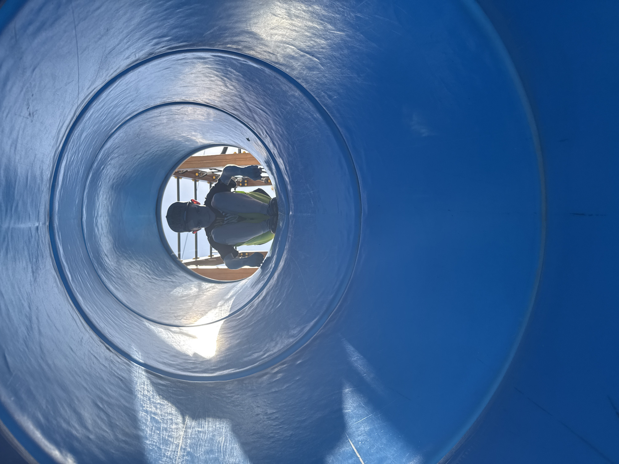 A person is seen at the end of a blue, circular tunnel. The person is wearing what appears to be dark clothing and light-colored shorts. The tunnel has concentric rings, and light is visible at the end. Part of a wooden structure is visible behind the person. The location is likely Shevlyakovo, Russia, though it is difficult to confirm with certainty from the provided image.