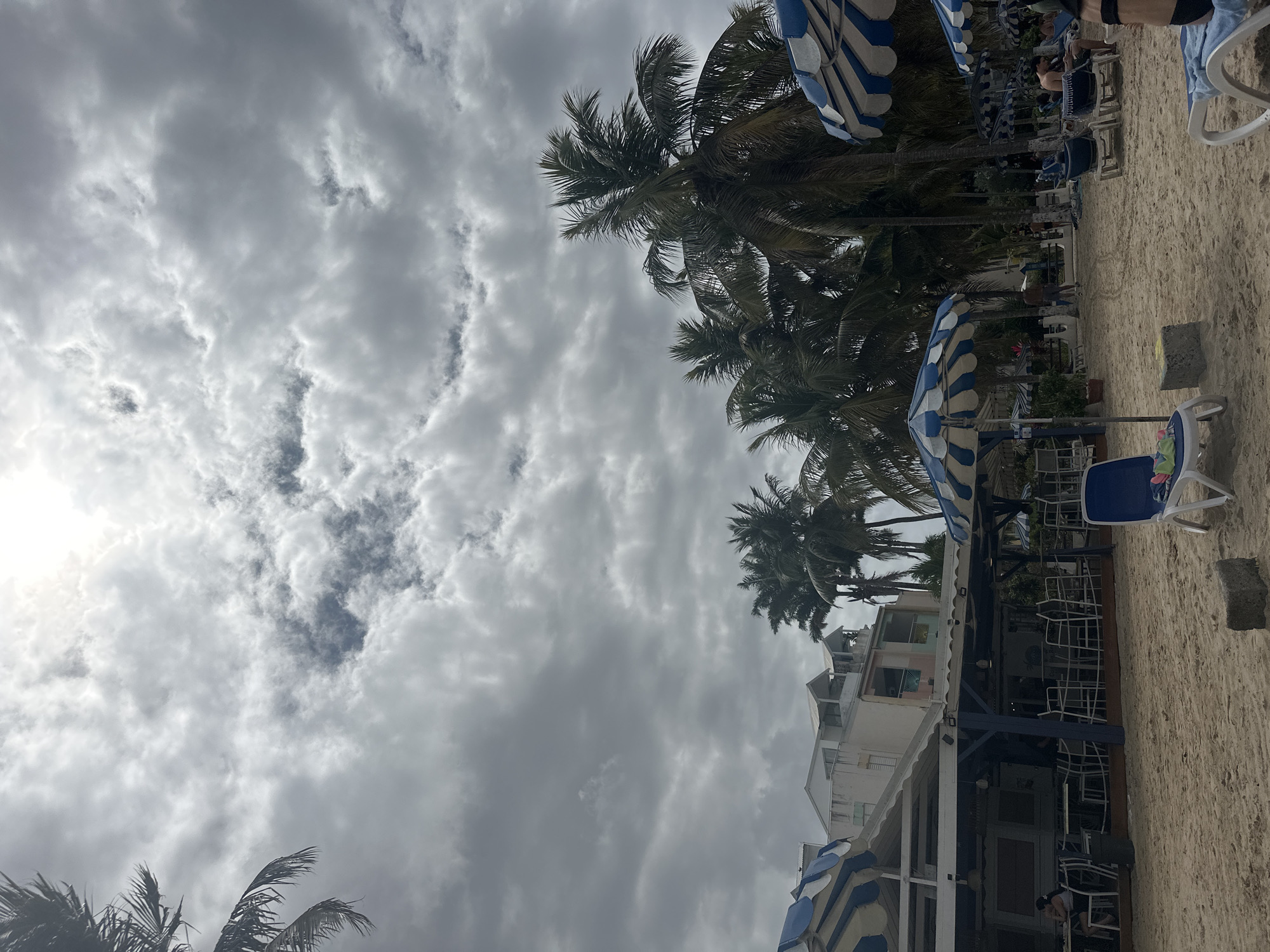 The image shows a beach scene, likely in Le Gosier, France, given the context. The sky is overcast with grey and white clouds dominating the upper left portion of the frame, while a few palm trees and several blue-and-white striped beach umbrellas are visible along the right side. A building with white and light pink walls is also present near the beach. Beach chairs and some people are seen near the umbrellas, indicating a relaxed beach atmosphere. The foreground shows sandy ground, likely the beach itself. The scene overall suggests a coastal area in a tourist location, possibly during cloudy weather.