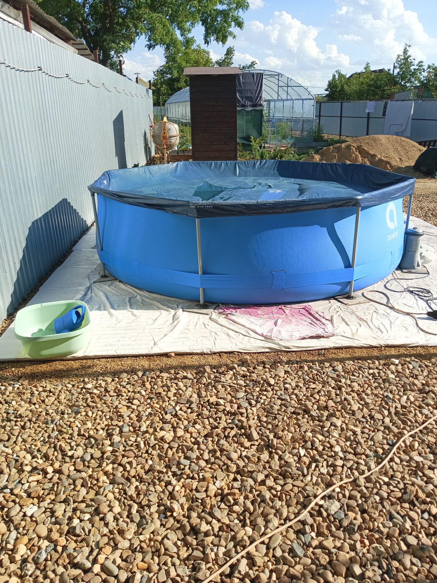 In the city of Maryanskaya, Russia, an outdoor scene depicts a large, blue, above-ground swimming pool with a dark blue cover. The pool is set up on a white tarpaulin spread over a gravel surface. A light green plastic washbasin containing a blue object, possibly a rolled-up mat or hose, is positioned to the left of the pool. In the background, a corrugated metal fence runs along the left side of the image. Further back, a wooden shed, a greenhouse structure, and piles of sand and gravel are visible. The sky above is blue with scattered white clouds. A power cord is visible on the gravel in the foreground, extending towards the right.