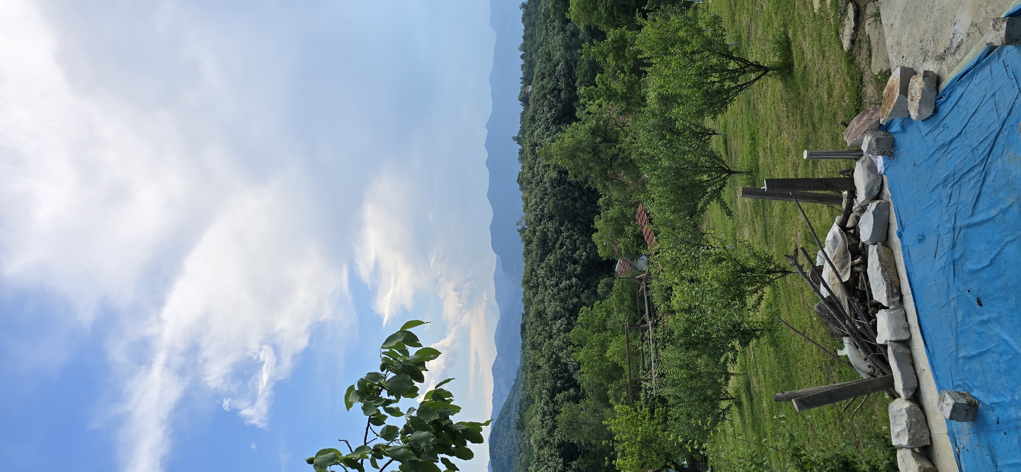 A vertical, outdoor, medium shot captures a panoramic view of a rural landscape under a partly cloudy sky. In the foreground, a blue tarp covers what appears to be a shallow pool or constructed area, bordered by large, light-colored stones and a pile of wood debris. A few wooden posts stand near the debris. Beyond this, a manicured lawn is dotted with fruit trees, and a rustic structure with a red-tiled roof is partially visible amidst the greenery. Further in the distance, a dense forest covers rolling hills, which transition into hazy blue mountains on the horizon. The sky above is a vibrant blue with scattered white clouds, some of which are dramatically illuminated by sunlight. A branch with green leaves and small green fruit hangs from the upper left corner, framing the scene. The overall impression is one of peaceful, rural tranquility.