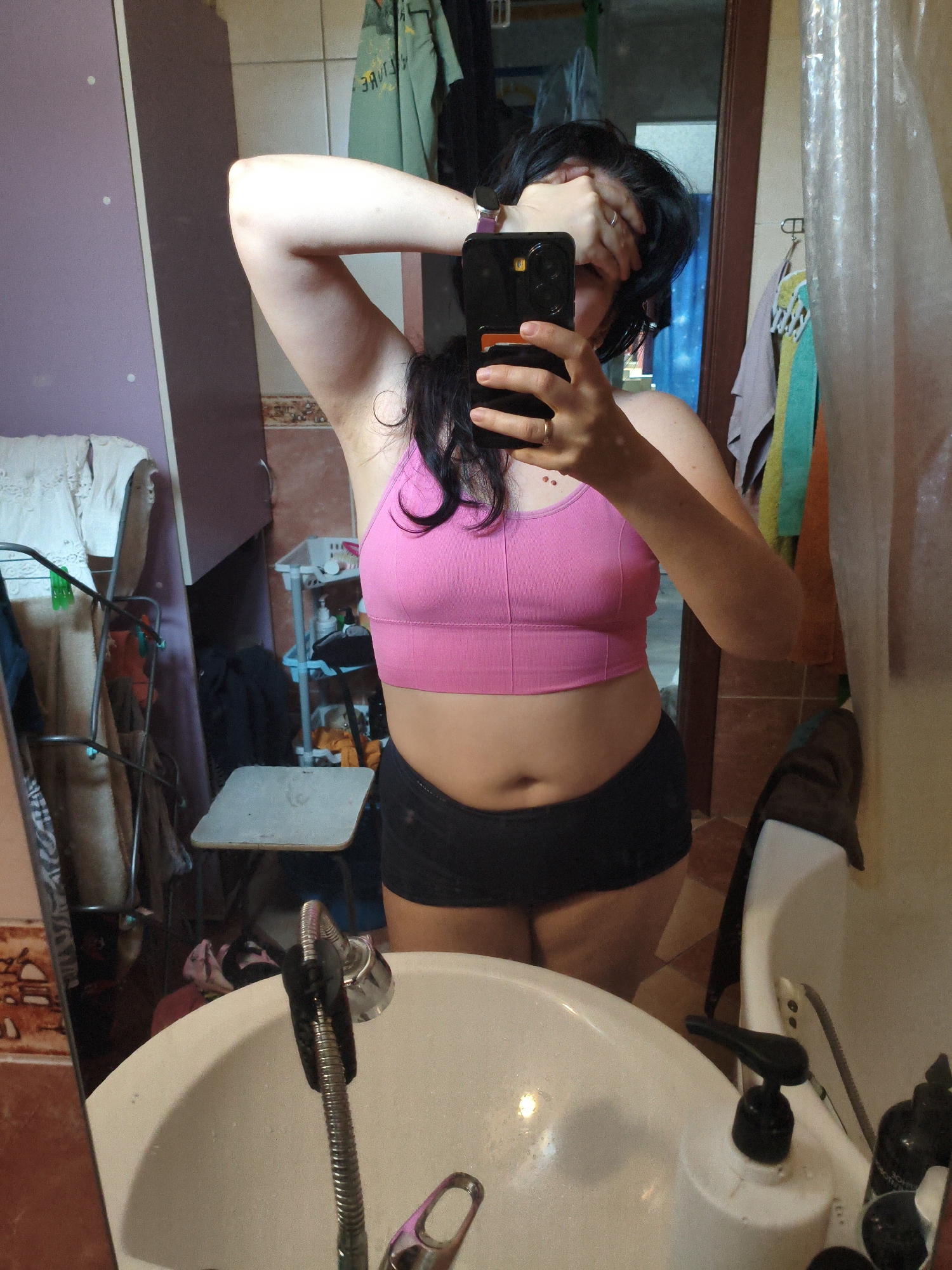 A full-body, selfie-style photograph captured in a bathroom. The subject, a person with dark hair, is standing in front of a mirror, holding a smartphone to take the picture. They are wearing a pink sports bra and black shorts. The bathroom appears cluttered with various items, including towels hanging on a rack, a shelf with toiletries, and a laundry basket. The sink and faucet are visible in the foreground, and a shower curtain is to the right. The overall impression is of a casual, everyday moment captured in a personal space. There is no discernible location in the city of Orenburg, Russia.