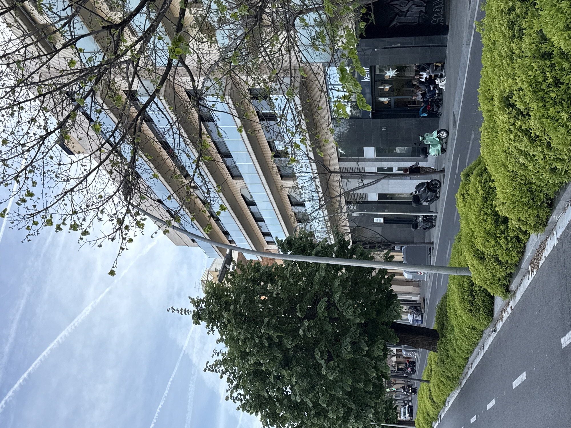 The image shows a street scene in Barcelona, Spain, featuring a modern building with reflective windows, a row of parked motorbikes, a tree-lined sidewalk, and a road with white lane markings. In front of the storefront at address 71, people are sitting at outdoor tables. Contrails are visible in the sky.