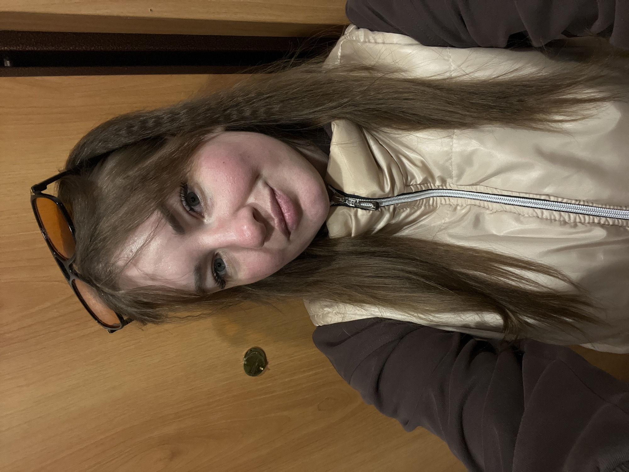 A young woman is posing indoors. She has long brown hair with bangs and is wearing a tan puffer vest over a brown long-sleeved shirt. She is also wearing brown-tinted sunglasses. The background appears to be a wooden door with a peephole. The photograph was taken indoors at an unknown location in the city of Chaikovsky, Russia.