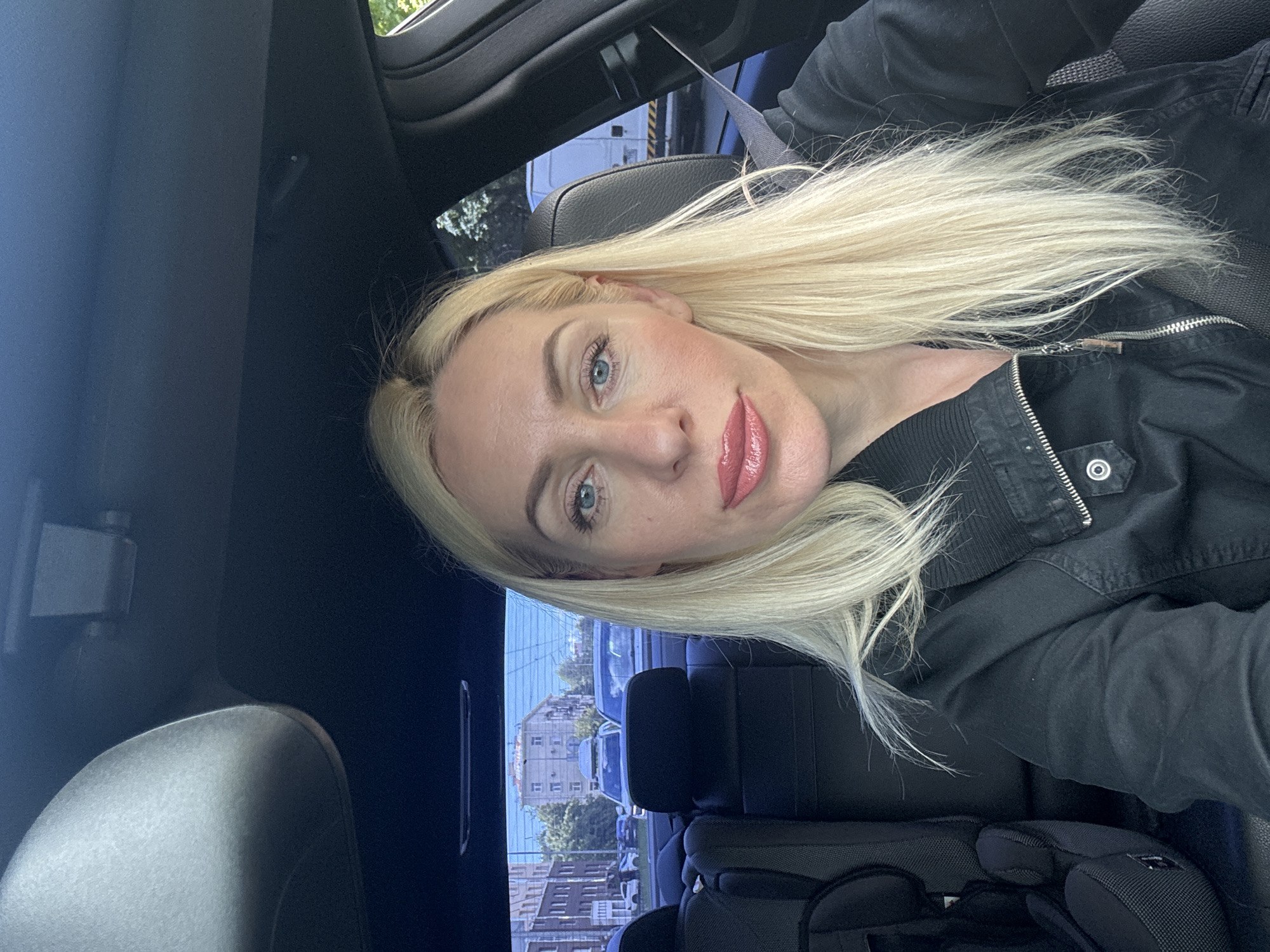 A blonde woman with blue eyes and pink lipstick is sitting in the driver's seat of a car. She is wearing a black jacket. The interior of the car is visible, including the headrest of the passenger seat and a portion of the back seat with a child car seat. Through the rear window, a city street scene with buildings and other cars can be seen. The lighting suggests it is daytime.