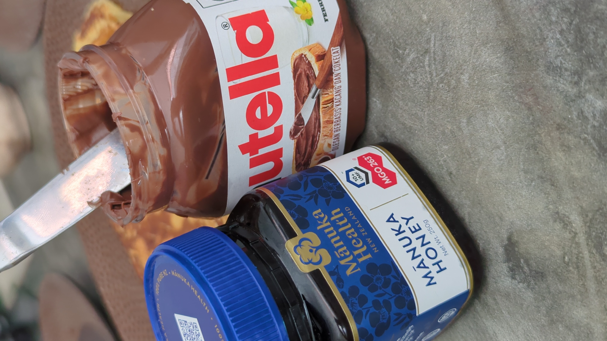 In North Kuta, Indonesia, on a textured gray surface, two jars are prominently displayed. To the left, a dark glass jar with a blue lid, labeled "Mānuka Health NEW ZEALAND," contains honey. The label features a floral pattern and text indicating "MĀNUKA HONEY Net Wt. 250g" and "MGO 263+". To the right, a larger jar of Nutella, partially opened, reveals a thick, chocolate spread. A silver knife with a wooden handle is inserted into the Nutella, with some spread on its blade. The Nutella jar's label prominently displays the brand name "Nutella" in red lettering. In the background, out of focus, appears to be a brown surface and a light-colored object, possibly food.