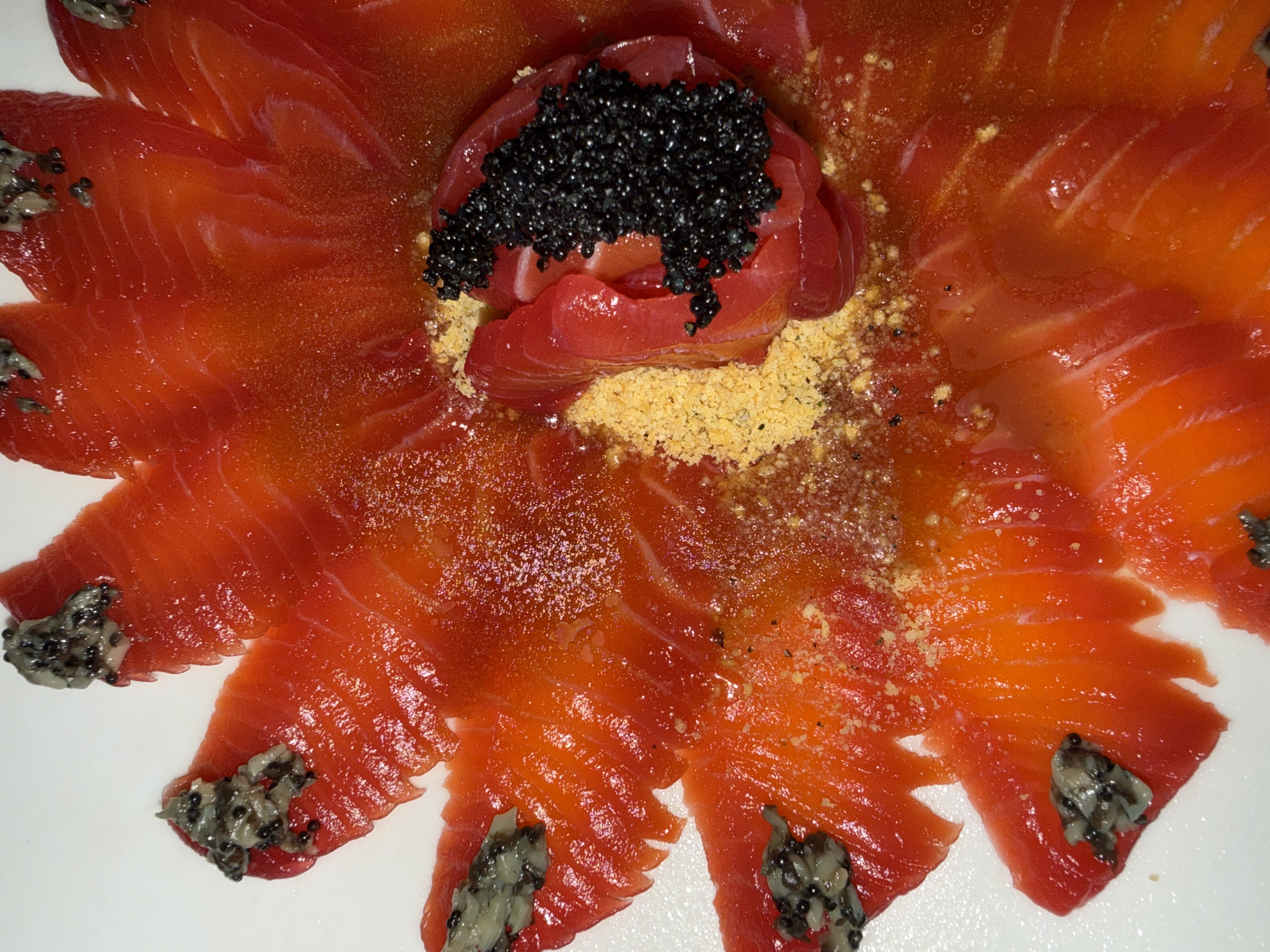 The image shows a plate of thinly sliced salmon arranged in a circular pattern. In the center is a mound of salmon topped with black caviar, sitting on a bed of yellow crumbs. The tips of the salmon slices are adorned with a grey, granular topping. The scene appears to be indoors, with a simple white background. The location is unspecified, and no people, actions or interactions are visible.