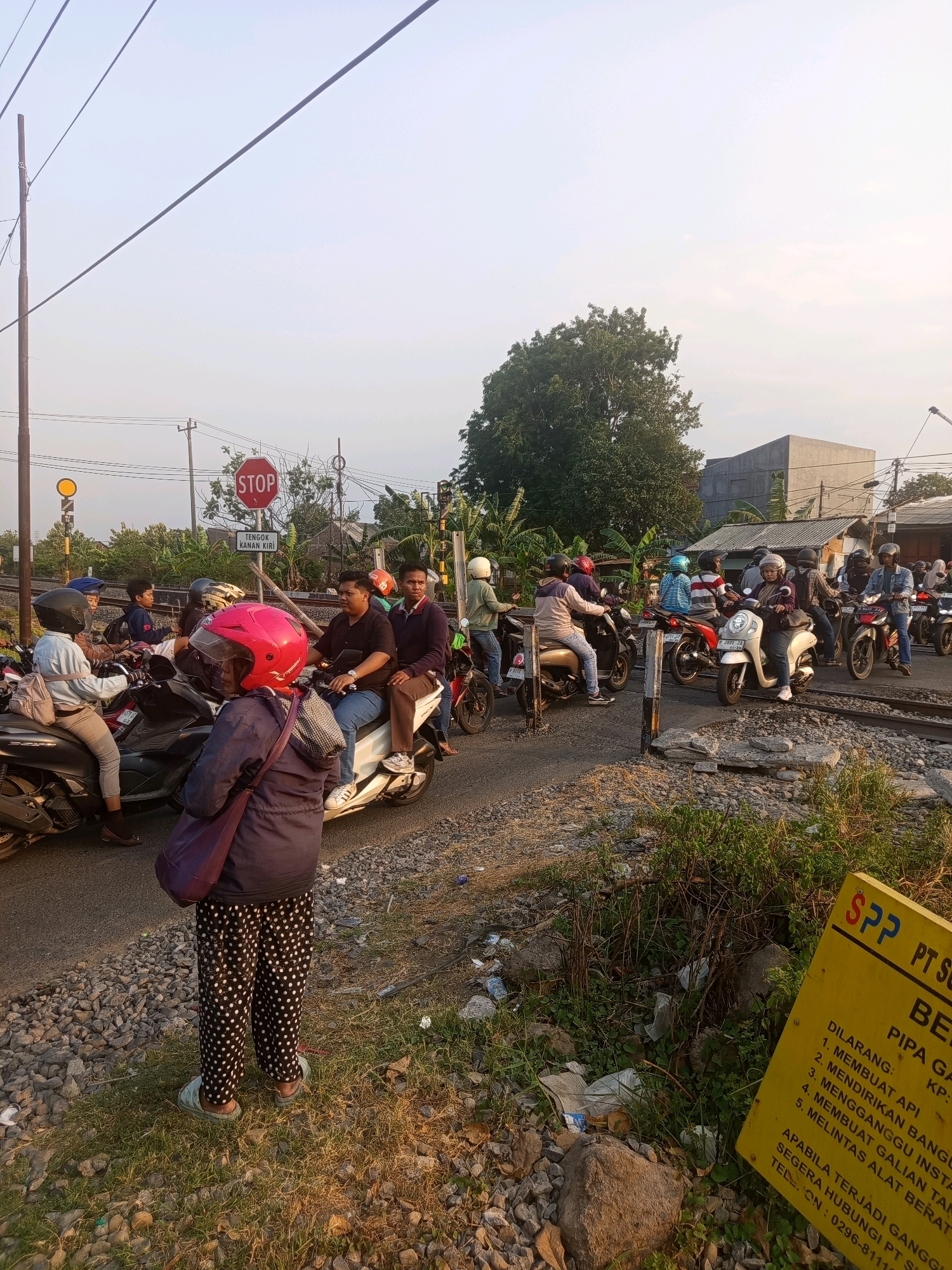 The image depicts a scene at a railway crossing in Semarang, Indonesia. Several motorcycles and their riders are stopped at the crossing, waiting for a train. A woman in a pink helmet and polka dot pants stands in the foreground, facing away from the camera. A yellow sign with red lettering is visible in the lower right corner, indicating regulations related to the railway crossing. The sky is clear and the sun is setting, casting a warm light on the scene. The environment appears to be a mixture of urban and rural elements, with buildings and trees visible in the background.
