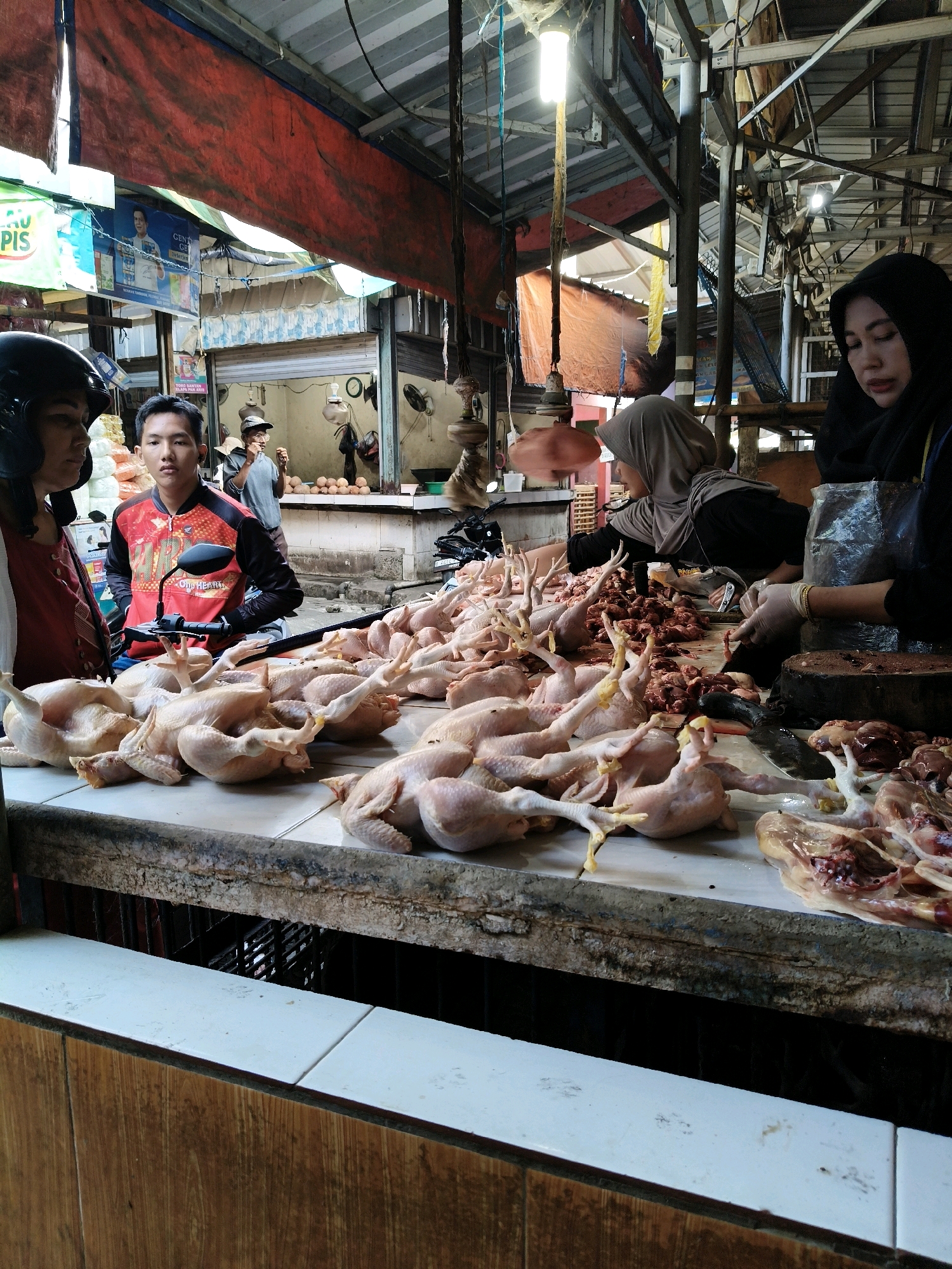 A medium shot captures a bustling outdoor market stall in Setiamekar, Indonesia, on a bright day. In the foreground, a long counter is laden with numerous raw chickens, some whole and others partially processed, arranged in rows. A pile of chicken innards is visible to the right.
Three individuals are present. On the left, a person wearing a black helmet and a red top looks towards the camera, partially obscured. To their right, a young man in a red and black patterned jacket sits on a motorcycle, facing forward with a neutral expression. To the right, two women are engaged in preparing the chickens. One, wearing a grey hijab, is focused on her work behind the counter. Another woman, veiled in black, is wearing gloves and appears to be actively cutting or handling the poultry with a knife. She is positioned in front of a large wooden chopping block, with her hands and forearms in view.
The market stall is covered by a corrugated metal roof supported by metal beams, with a large red tarpaulin hanging in the background. Various signs and posters are visible on the walls behind the counter, including what appears to be an advertisement. The overall environment suggests a busy and active marketplace.