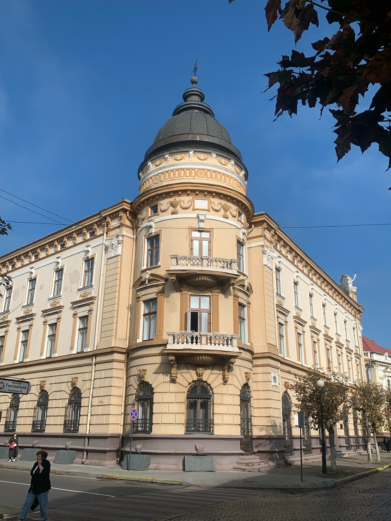 The image captures a corner of a cream-colored building with a dome and a balcony, situated in the city of Kolomyia, Ukraine, on a clear day. The architectural style appears to be ornate, with detailed window frames and decorative elements along the facade. A street runs in front of the building, paved with cobblestones. A pedestrian crossing is visible. Two individuals are present: one woman is walking across the street, and another is standing on the sidewalk, engaged in a phone conversation. Trees with autumn foliage line the street, and there are overhead power lines. A street sign in Ukrainian, indicating "Teatralna St.", is also visible. The sky is a vibrant blue, with some tree branches extending into the frame from the upper right corner.