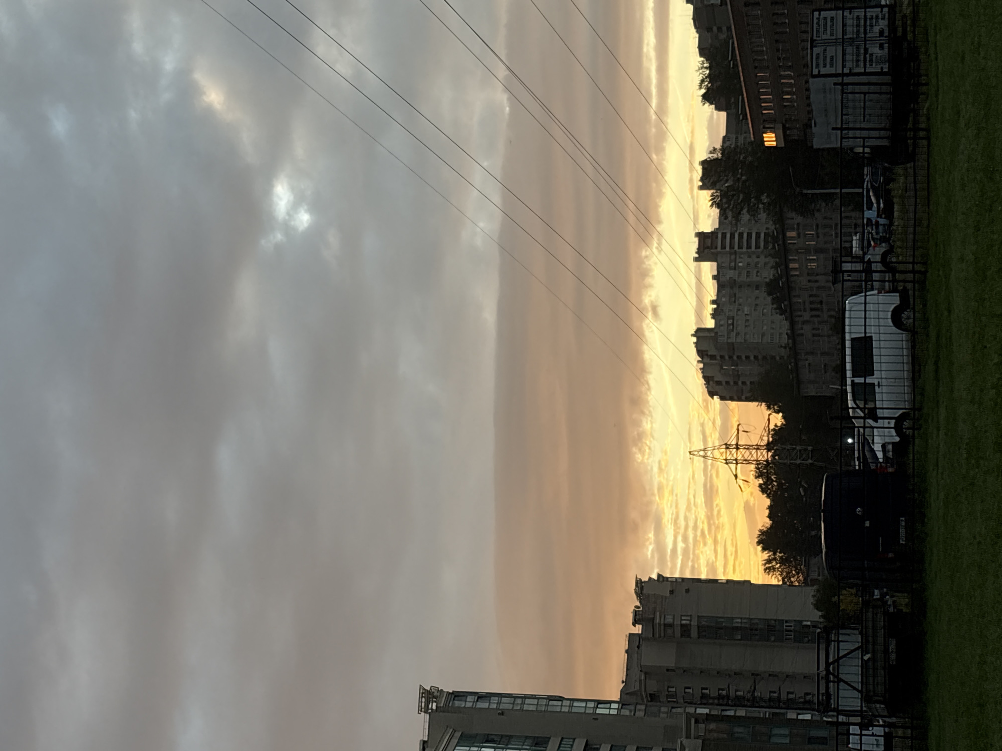 The image depicts a street scene in Saint Petersburg, Russia, during a cloudy sunset. The sky is filled with a mix of gray and orange-hued clouds, with a brighter patch of sunlight breaking through in the distance. Several tall residential buildings, typical of Soviet-era architecture, are visible on the left and in the background. Power lines crisscross the sky diagonally from the upper right to the lower left. In the foreground, there is a grassy area bordered by a dark fence. Behind the fence, several vehicles are parked, including a black van, a white minivan, and a flatbed truck. A large electricity pylon stands prominently behind a row of trees. The overall impression is one of an urban environment at dusk, with the fading daylight casting a warm glow on the buildings.