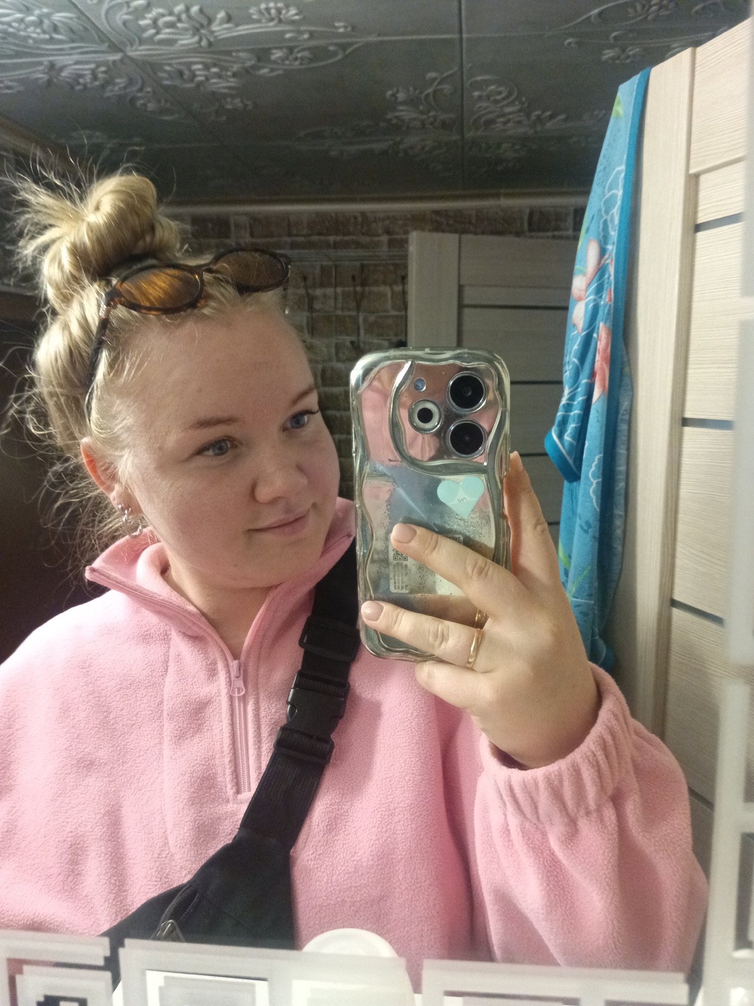 A woman is taking a selfie in what appears to be a bathroom. She has her blonde hair in a messy bun with sunglasses on top of her head. She is wearing a pink fleece zip-up sweater and a black fanny pack across her chest. She holds an iPhone with a clear case, adorned with a small heart sticker, which is reflecting the bathroom environment. A gold ring is visible on her ring finger. The background includes a textured ceiling, brick-like wall, a partly visible blue patterned towel hanging on a light-colored door, and the reflection of the bottom edge of a mirror with small decorative frames. The location is assumed to be in the city of Фоки, Russia.