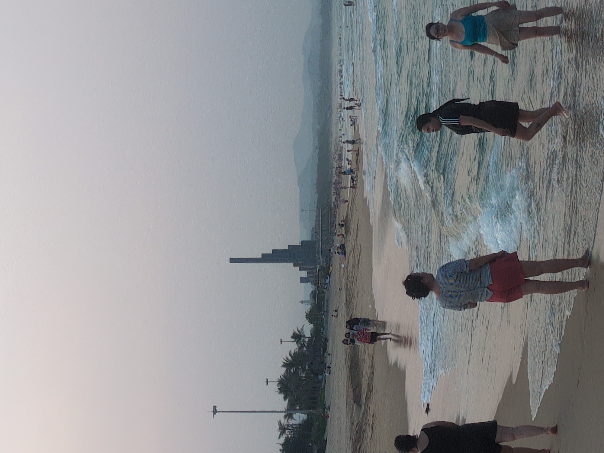 The image captures a beach scene in Tuy Hòa, Vietnam. The beach is populated with people wading in the surf and walking along the sand. A stepped high-rise building is visible along the coastline. Palm trees and light posts are present along the edge of the beach. The sky is hazy.