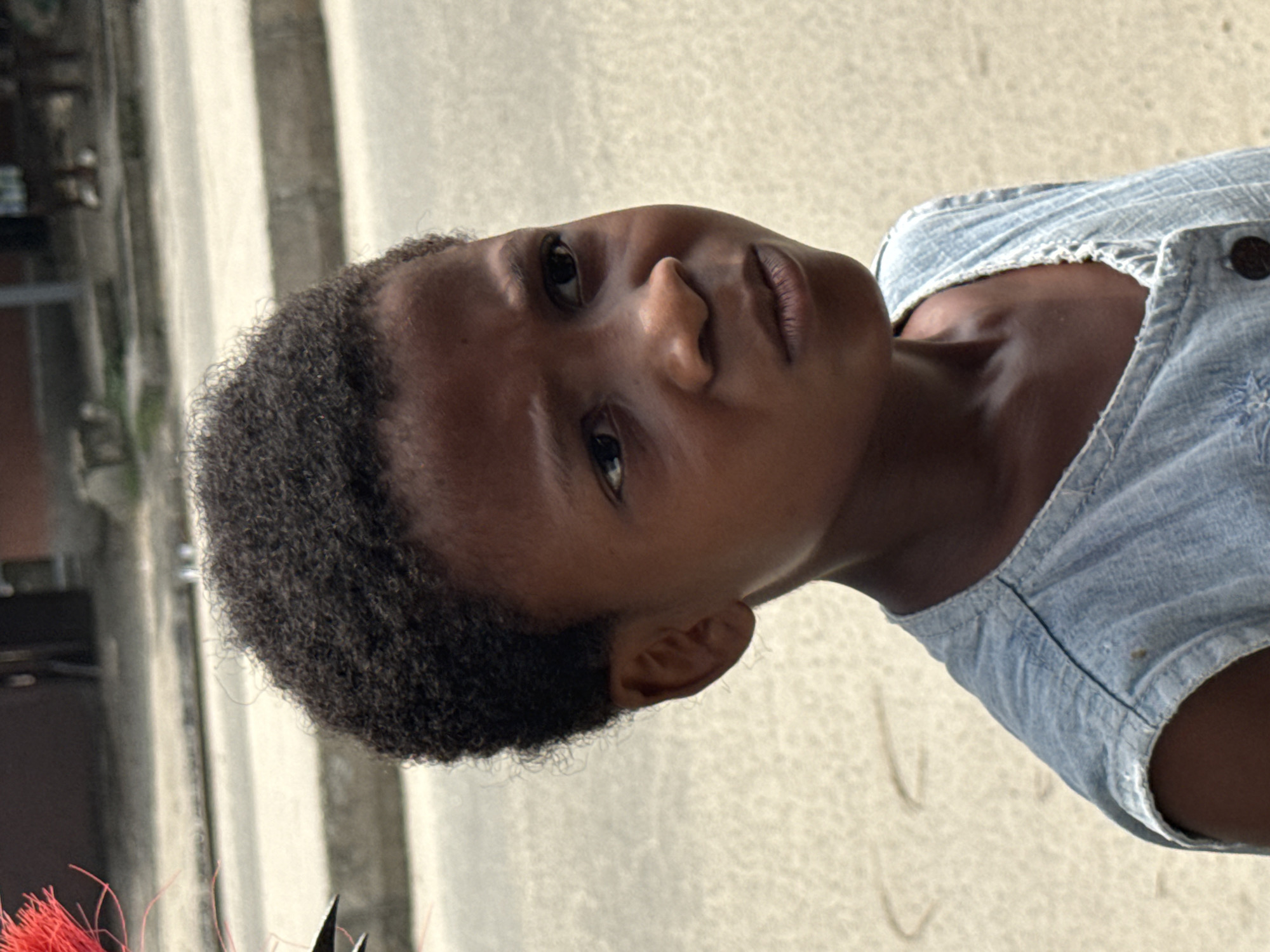 In the city of Ado, Nigeria, a young person with dark, curly hair is depicted from the chest up, facing the viewer's right. They are wearing a light blue denim sleeveless top with visible stitching and a dark button. Their expression is serious, with furrowed brows and a slightly downturned mouth. The background is out of focus, showing a paved surface that appears to be a street or walkway, with concrete structures and glimpses of other people in the distance. Some red feathery material is visible in the upper left corner of the frame, partially obscuring the subject's head.