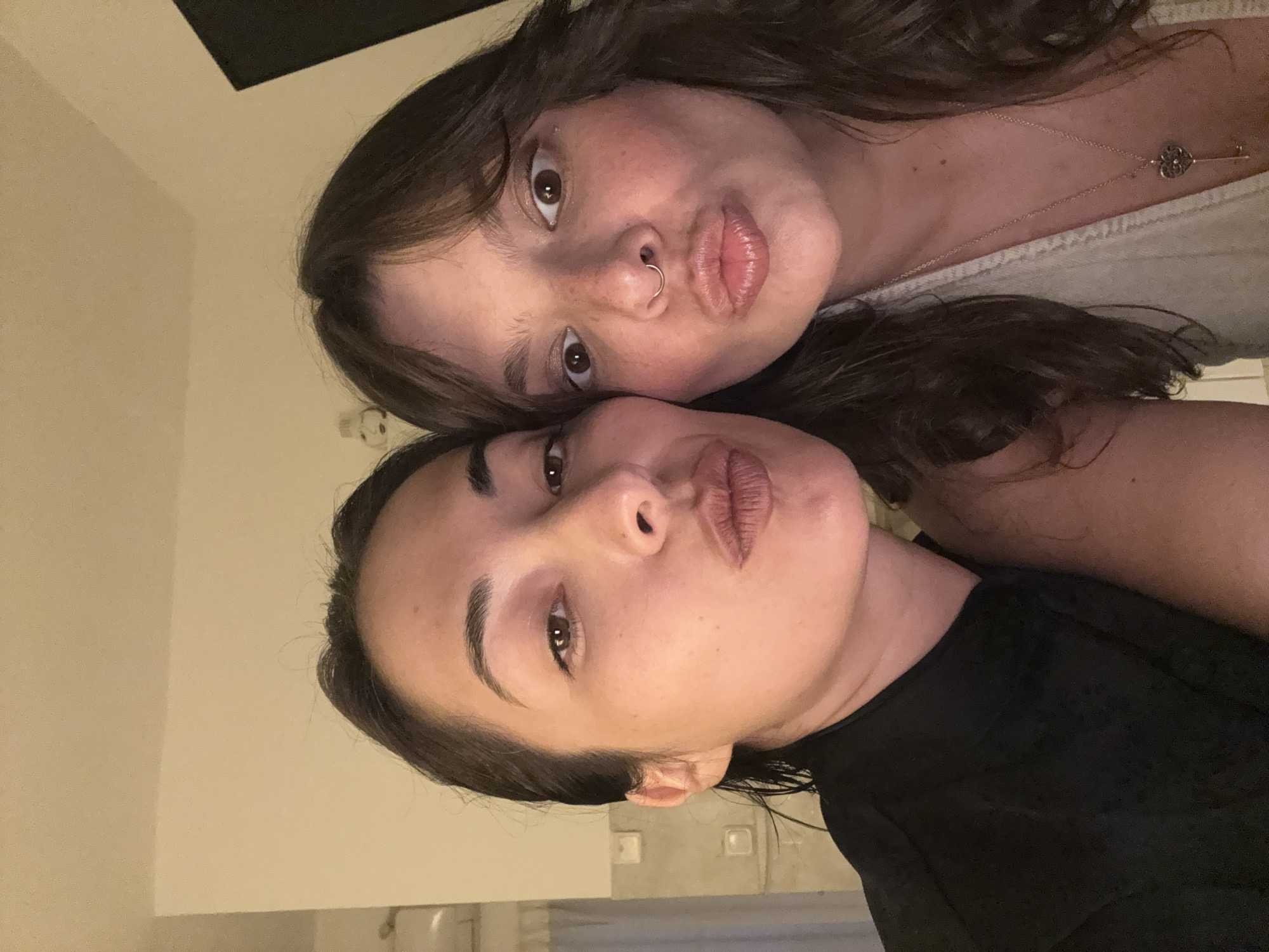 Two women are posing closely together indoors. The woman on top has medium-length brown hair, a nose ring, and is wearing a light-colored top and a necklace with a heart-shaped pendant. The woman below her has short, dark hair and is wearing a black top. Both women are making a kissing face. The background is a plain wall, with hints of light fixtures suggesting an indoor environment. The location cannot be determined to be in Tel-Aviv, Israel, as there are no visible features in the photo to suggest this.