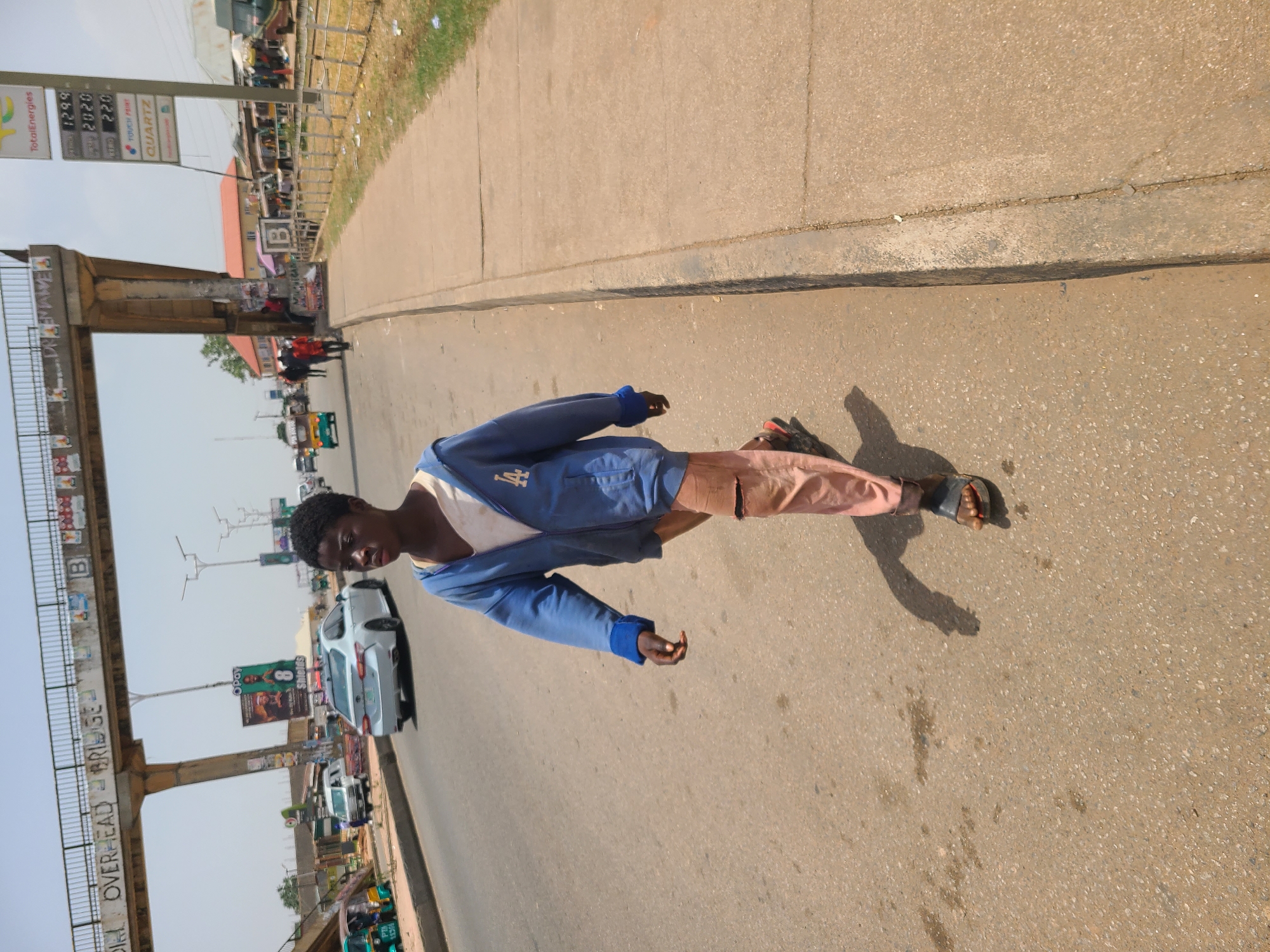 The image shows a young man standing on a concrete surface, likely a sidewalk or the edge of a road. He is wearing a blue jacket with a white shirt underneath, light brown pants, and sandals. His shadow is cast on the ground beside him.
In the background, there is a busy street scene with cars, motorcycles, and pedestrians. An overhead bridge or structure is visible, with the words "OVERHEAD BRIDGE" clearly readable. There is also a gas station sign in the background displaying fuel prices and brands. The overall environment appears to be a typical urban setting in Jos, Nigeria. The street scene and the gas station are indicative of a roadside environment, where people are buying and selling.