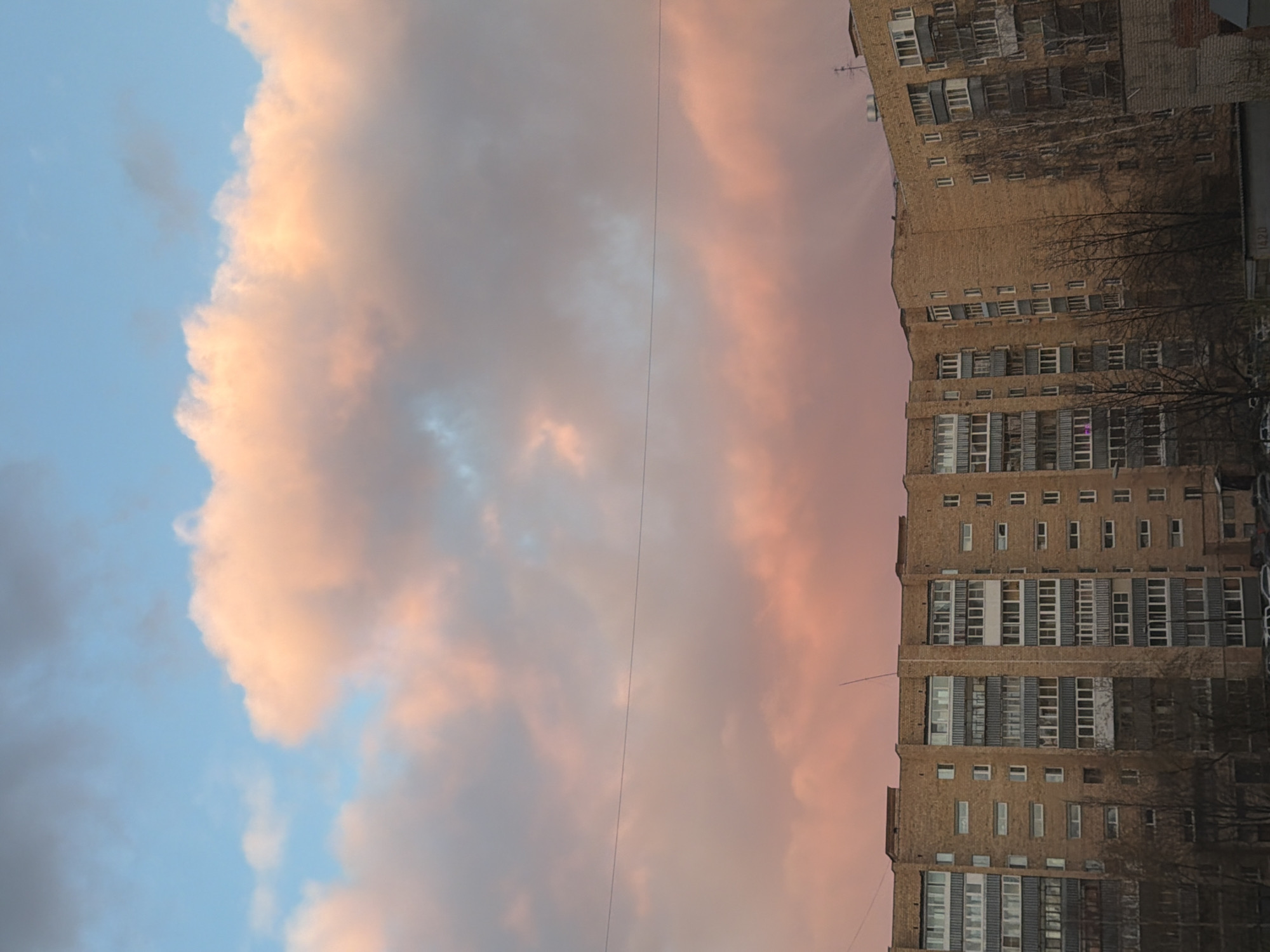 The image depicts a high-rise brick building on the right side of the frame and a cloudy sky on the left. The sky is colored with hues of pink, gray, and blue. A power line stretches horizontally across the middle. The building features rows of windows, and bare trees are visible near its base. The location is likely Yekaterinburg, Russia.
