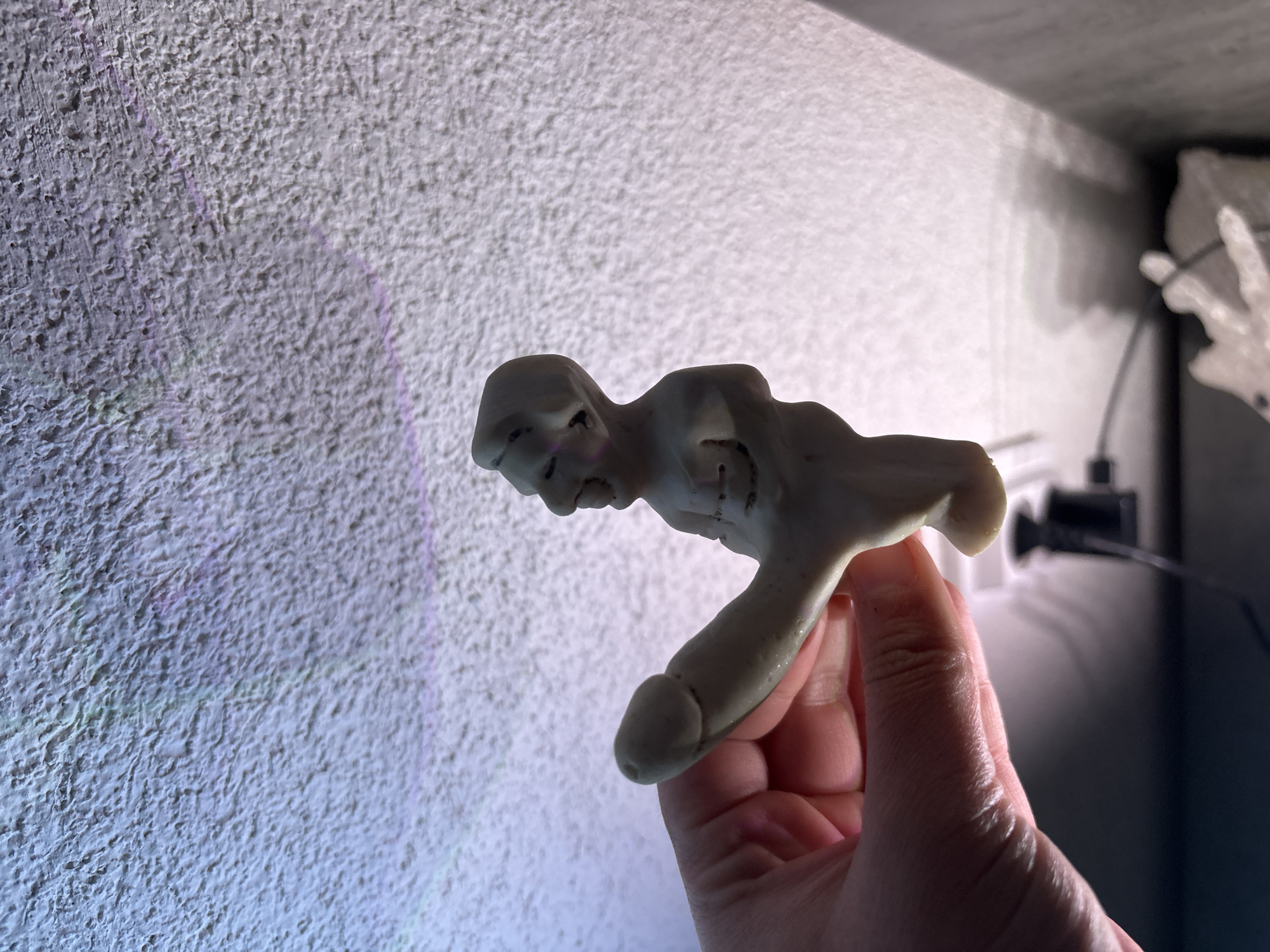 A hand is holding a small, misshapen, white figurine against a textured white wall. The figurine appears to be vaguely human-shaped with a distorted face and body. The background is a combination of bright white wall and a darker area in the upper right containing a black object with a cord. The image was taken indoors with the hand appearing to be in focus. The image location, city of Kuklen, Bulgaria, is not apparent in this scene.