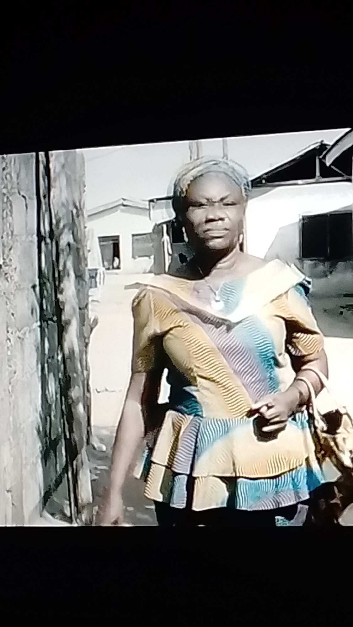 In Lokoja, Nigeria, a woman stands outdoors, facing the camera. She has dark skin, gray hair wrapped in a light-colored headscarf, and is wearing a patterned blouse with a ruffled hem, featuring diagonal stripes in yellow, blue, and white. She is adorned with earrings and bangles on her left wrist. In her right hand, she holds a small object, possibly a phone, and her left hand rests on a woven bag slung over her shoulder. The background shows a narrow alleyway with a textured concrete wall on the left and houses with open doorways and windows on the right. The overall lighting suggests it is daytime.