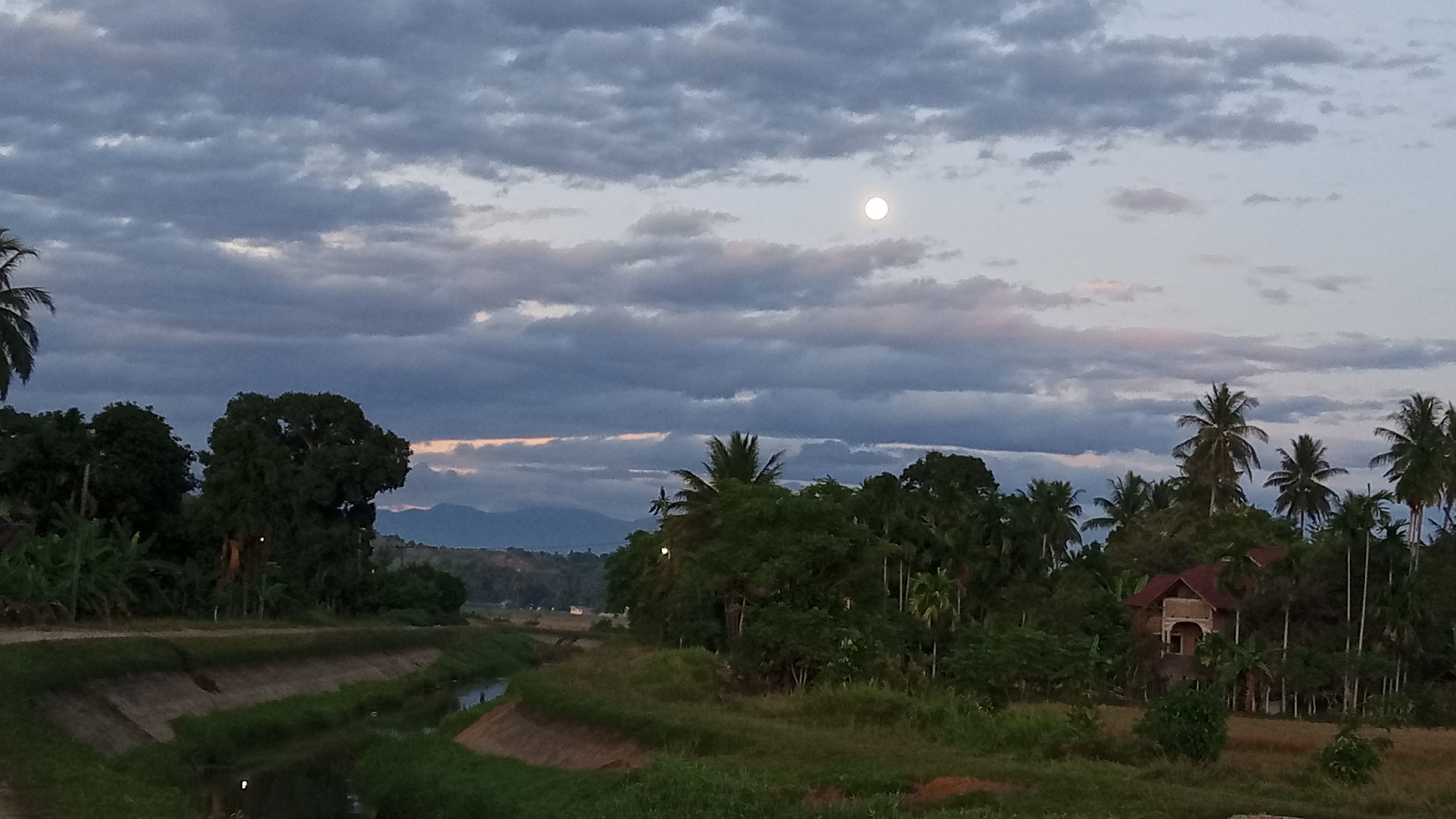 The image depicts a tranquil rural landscape at dusk, likely in Buketteukuh, Indonesia. A full moon shines brightly in the twilight sky, which is adorned with dramatic, textured clouds. The foreground features a man-made canal with concrete banks, flanked by lush green grass and vegetation. To the right of the canal, a cluster of palm trees and dense foliage dominate the scene, with a rustic-looking house nestled amongst them. In the distance, rolling hills and mountains fade into the hazy horizon. The overall atmosphere is serene and peaceful, capturing a moment of natural beauty as day transitions into night.