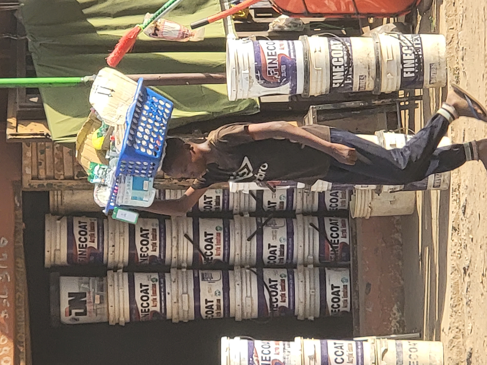A man balances a blue plastic basket on his head while lying on his side atop stacks of "FINECOAT" and "NECOAT" paint buckets in an outdoor setting. The location is suggested to be Maiburiji, Nigeria based on the context. The man is wearing a dark shirt, dark pants, and sandals. Above him and to the side are various items on some sort of rack. The paint buckets are stacked in an orderly fashion. The scene is likely captured in daylight.