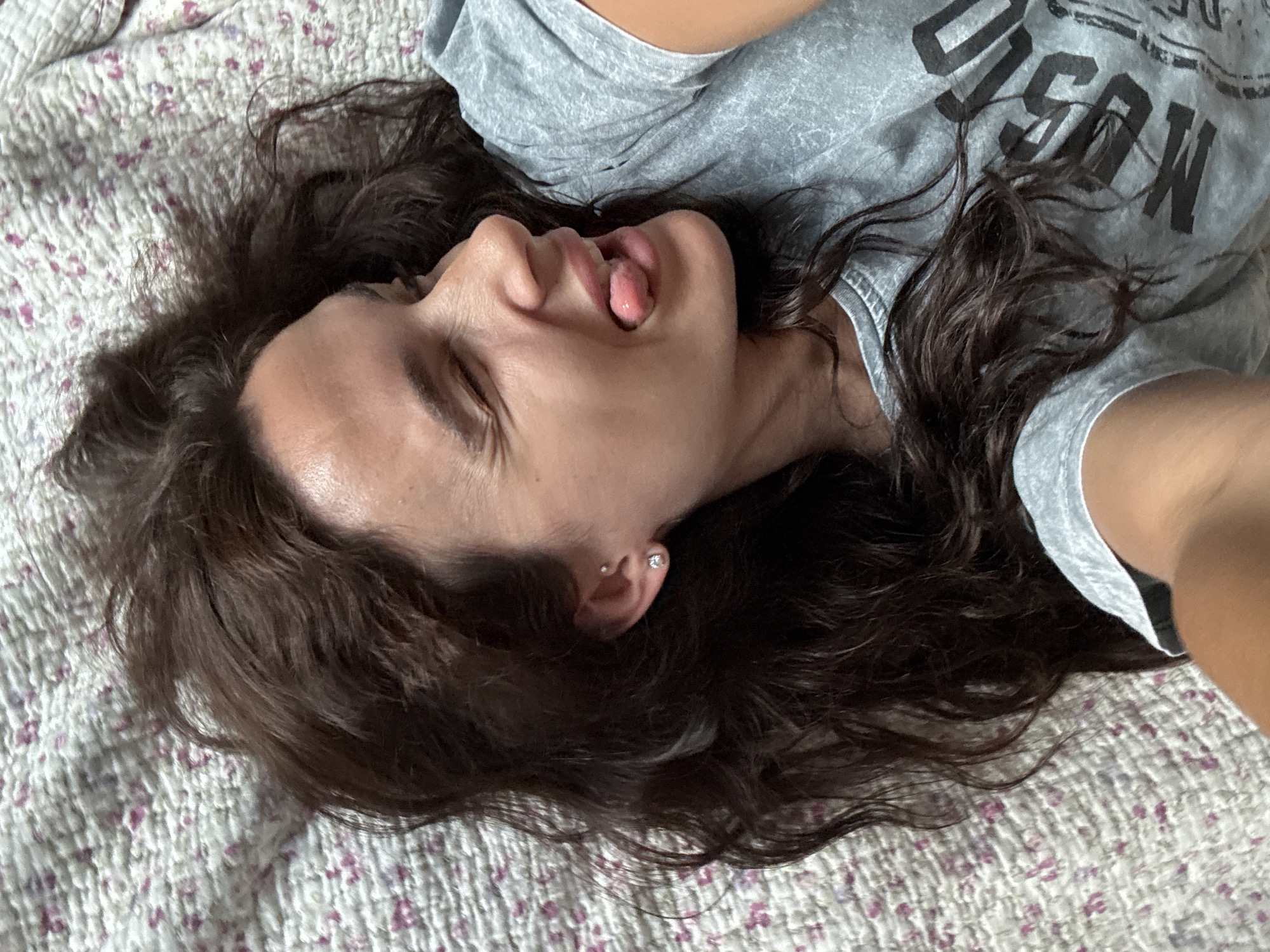 A medium shot, taken from above, shows a woman lying on her side on a floral patterned quilted bedspread. Her dark brown hair is spread out around her head. Her eyes are closed, and her right cheek is pressed against the bedspread. Her mouth is open, and her tongue is sticking out. She is wearing a grey t-shirt with black lettering on it. The background is the floral patterned bedspread, with a mix of pink and white flowers. The image does not contain any information that would allow for a determination of its location in Chaikovsky, Russia.