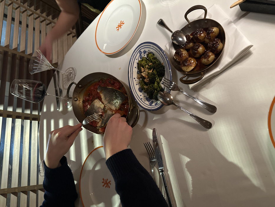 A table setting displays plates with orange trim, one empty and one partially obscured by an arm. A pot of fish in tomato sauce is being eaten with a fork. Additional dishes include broccoli with almonds on a blue-patterned plate and glazed potatoes in a small pan. Silverware is arranged near the plates, and two wine glasses are near the fish. Light and shadow play across the white tablecloth. A partial view of a person's dark blue sleeves is visible.