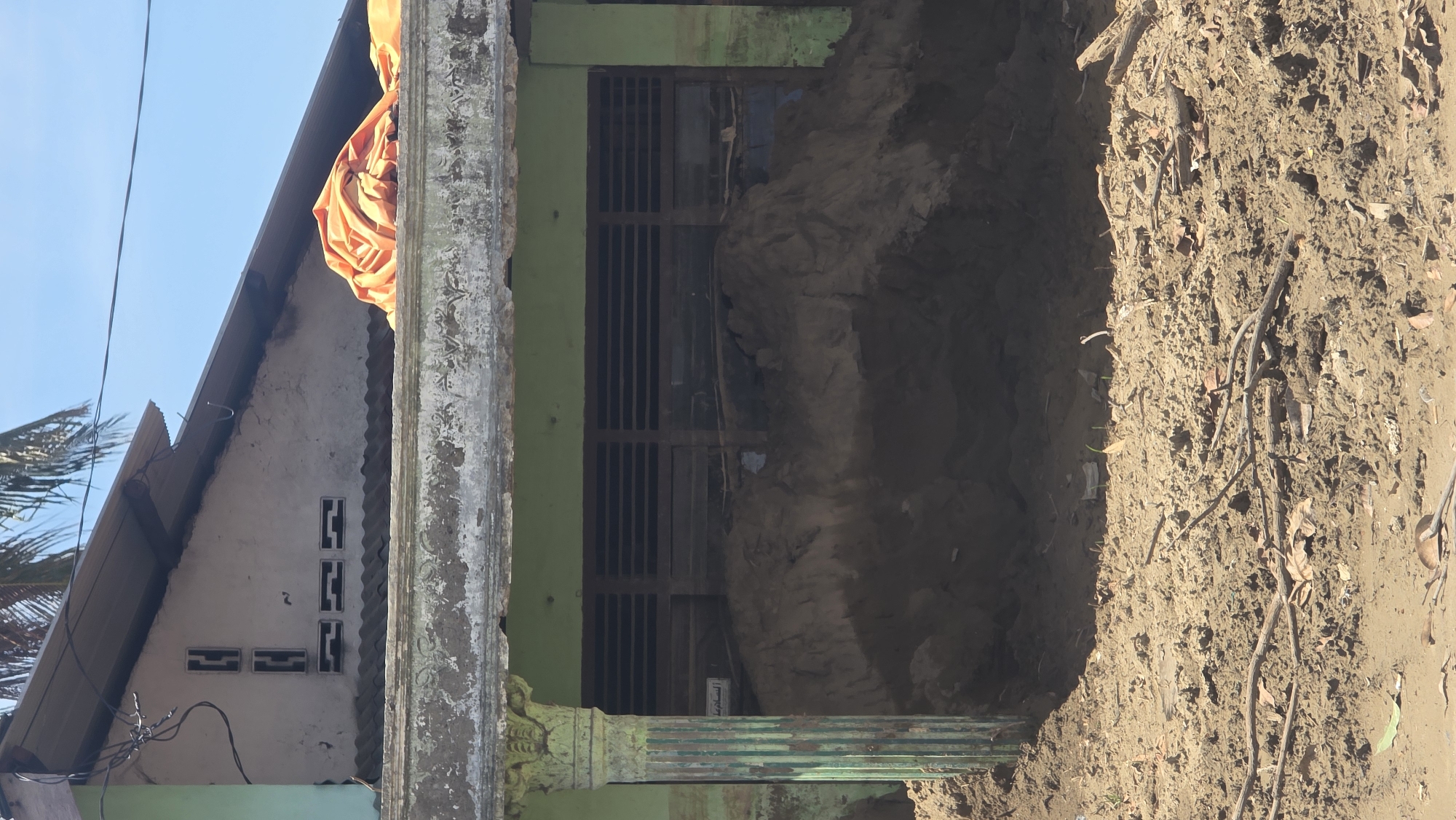 The image depicts a collapsed area next to a building in Manyang Cut, Indonesia. The structure, likely a house, has green trim around a window with horizontal bars. A beige cloth is draped over the edge of the roof of an adjacent structure. The ground on the right side of the building is a mix of dirt, leaves, and small branches, suggesting recent digging or a landslide. The area immediately next to the building has collapsed, creating a large hole exposing the earth underneath the structure.