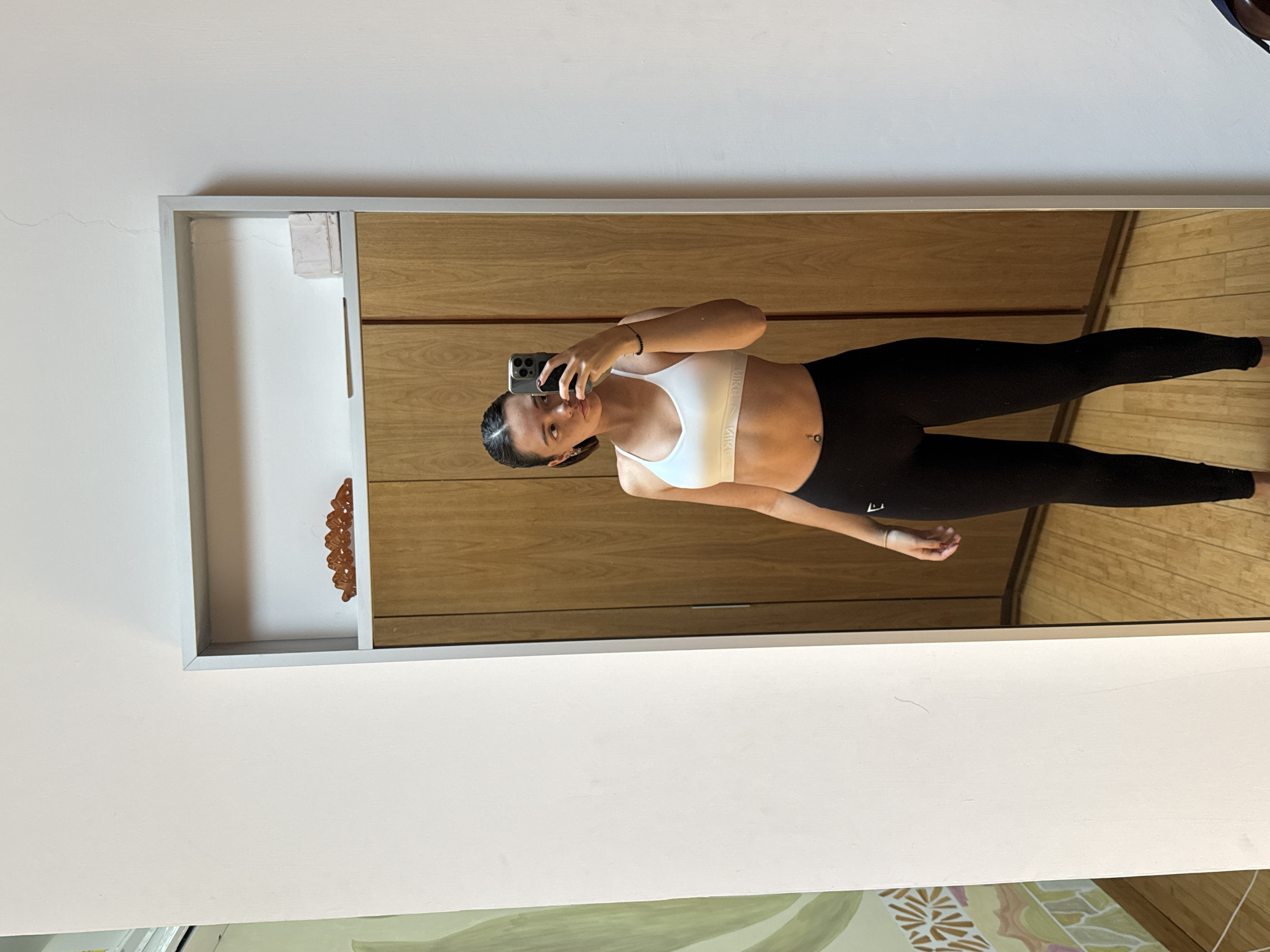 A woman in a white sports bra and black leggings is taking a selfie in a mirror. The mirror is rectangular with a silver frame and is mounted on a wall with a wooden cabinet behind it. The wall is white. The location is San Bernardino Tlaxcalancingo, Mexico.