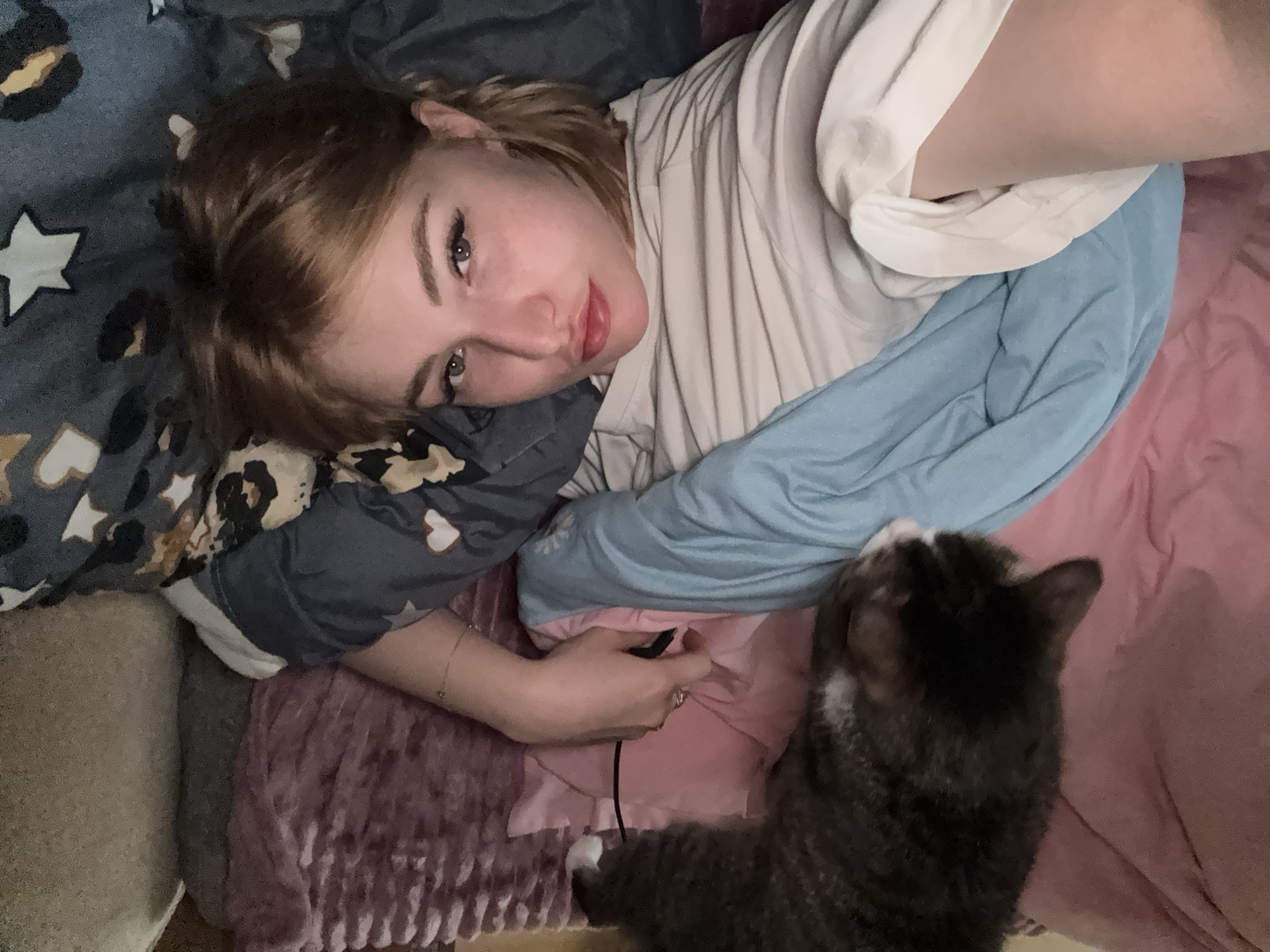 A woman with light brown hair and blue eyes lies in a bed with a cat. The woman is wearing a light-colored t-shirt and her makeup includes mascara and red lipstick. She has rings on her fingers and a bracelet on her wrist. The cat is a tabby with grey, black, and white fur and it is lying on top of the woman's arm. The bed is covered in blankets and pillows of various colors, including pink, blue, and purple. The scene appears to be indoors, possibly in a bedroom. There is no visual information in the image that can determine the location in the city of Khimki, Russia.