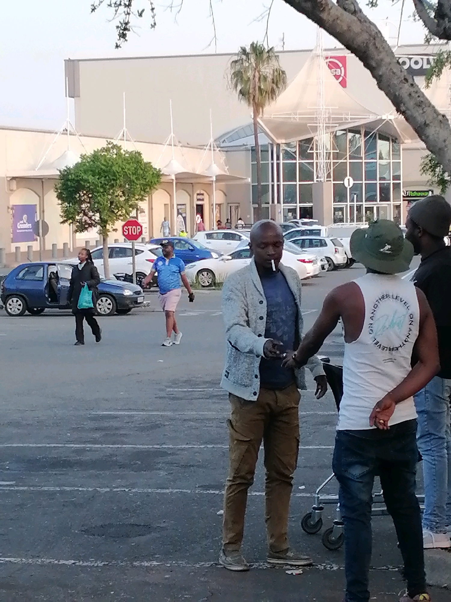 In a parking lot outside a shopping center, a group of men are gathered. One man, wearing a grey cardigan and brown pants, holds a cigarette in his mouth. He is facing another man in a white tank top with a circular graphic on the back and a green hat. To the right of this pair stands a third man in a black t-shirt and blue jeans. In the background, several cars are parked, and a blue hatchback is visible to the left. A woman in a black coat and a blue shopping bag is walking away from the blue hatchback. The shopping center features a large white awning over its entrance. The scene appears to be in the city of Emfuleni Local Municipality, South Africa.