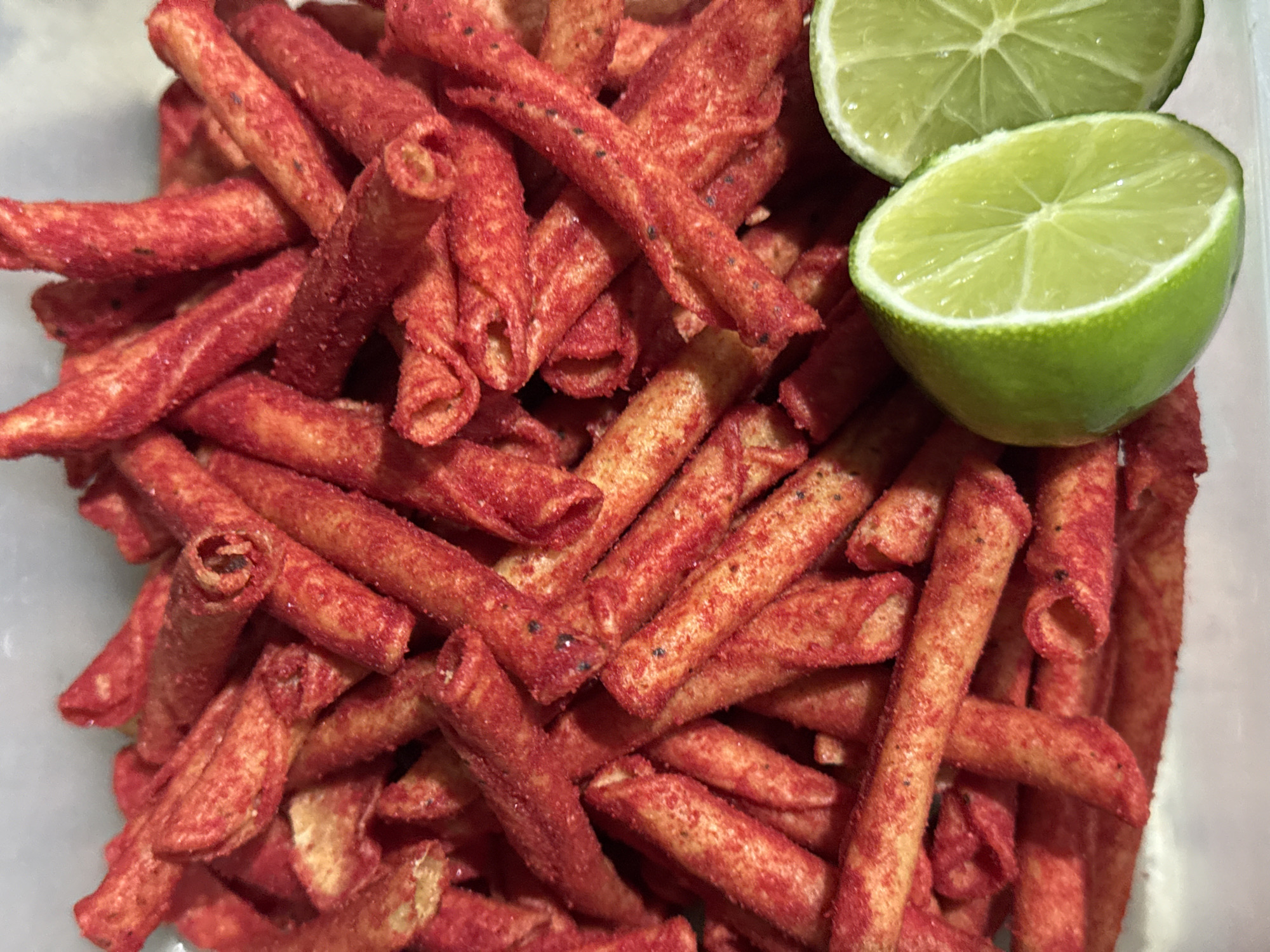 A close-up, high-angle shot captures a generous pile of red, rolled corn chips, likely Takis, within a clear or translucent container. The chips are uniformly cylindrical, covered in a vivid red seasoning with visible darker specks, suggesting a chili and spice coating. Their texture appears rough and crunchy from the seasoning.
To the right side of the frame, two halves of a fresh, bright green lime are prominently displayed. The limes are neatly cut, revealing their pale green, segmented flesh and juicy interior. The vibrant green of the limes creates a striking color contrast against the intense red of the chips.
The scene focuses solely on these food items, suggesting a snack ready for consumption. No activity is explicitly taking place. The lighting is even and soft, indicating an indoor setting. There are no people visible, nor any discernible text. The arrangement of the snack and the fresh lime is typical of how such items are enjoyed, particularly in regions like San Bernardino Tlaxcalancingo, Mexico, where chili-lime flavored snacks are popular.