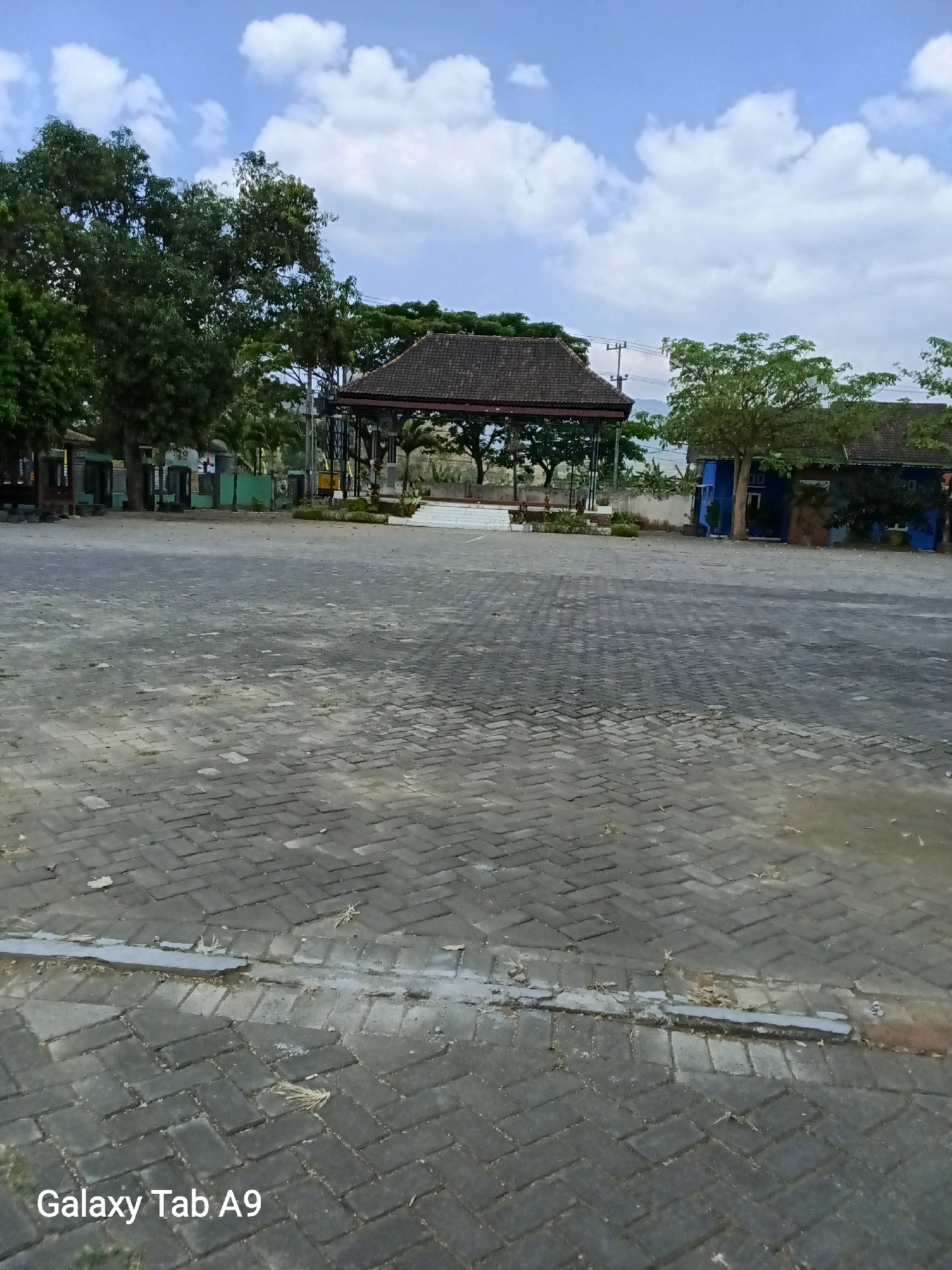 The image depicts an outdoor scene with a paved area in the foreground and a covered structure with a tiled roof in the background. The paved area is constructed from interlocking grey rectangular bricks arranged in a herringbone pattern. Small amounts of debris, including dry leaves and a light-colored strip of material, are visible on the pavement.
In the background, a gazebo-like structure with a dark, multi-faceted tiled roof stands on a raised platform. The structure has open sides supported by pillars and appears to be a public resting area or pavilion. Surrounding the pavilion are lush green trees with dense foliage. To the right of the pavilion, a blue building with a dark roof and visible windows is present. Power lines and poles are also visible in the distance.
The sky above is blue with scattered white clouds. The overall environment suggests a well-maintained outdoor public space, possibly a park or a communal gathering area in Karangan, Indonesia, given the watermark "Galaxy Tab A9" which is a Samsung tablet model often used in that region. There are no people visible in the image.