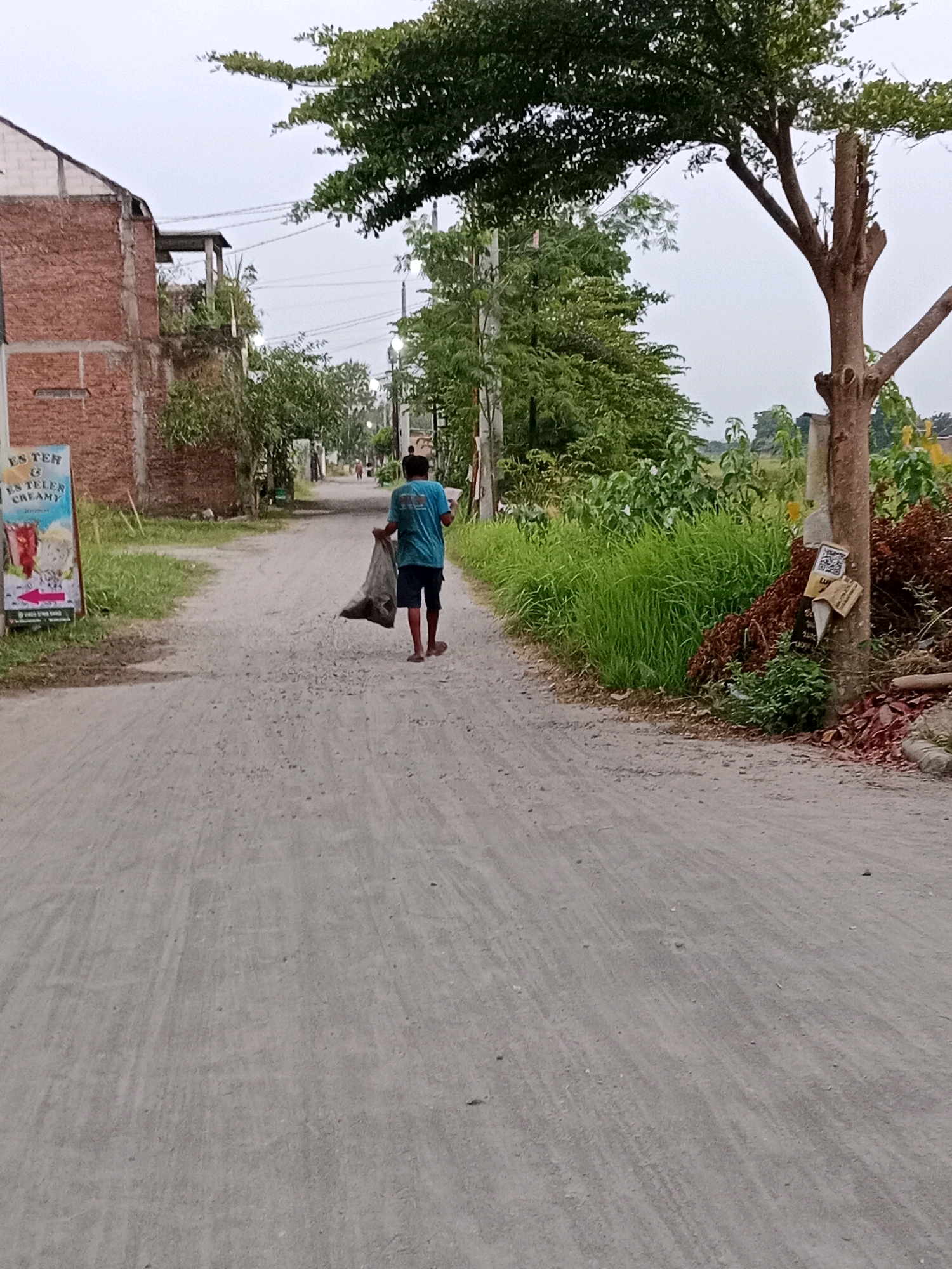 In Waru, Indonesia, a person walks down a dusty, unpaved road, carrying a dark plastic bag filled with debris. The individual, wearing a blue t-shirt and dark shorts, is seen from behind, moving away from the viewer. To the left of the road, a two-story brick building with exposed concrete supports is partially visible, adorned with a sign advertising "ES TEH & ES TELLER CREAMY." Lush green vegetation lines the right side of the road, with tall grass and trees creating a natural border. The sky is overcast, suggesting a subdued or early morning light. The overall scene depicts a quiet, rural or semi-rural street in Waru.