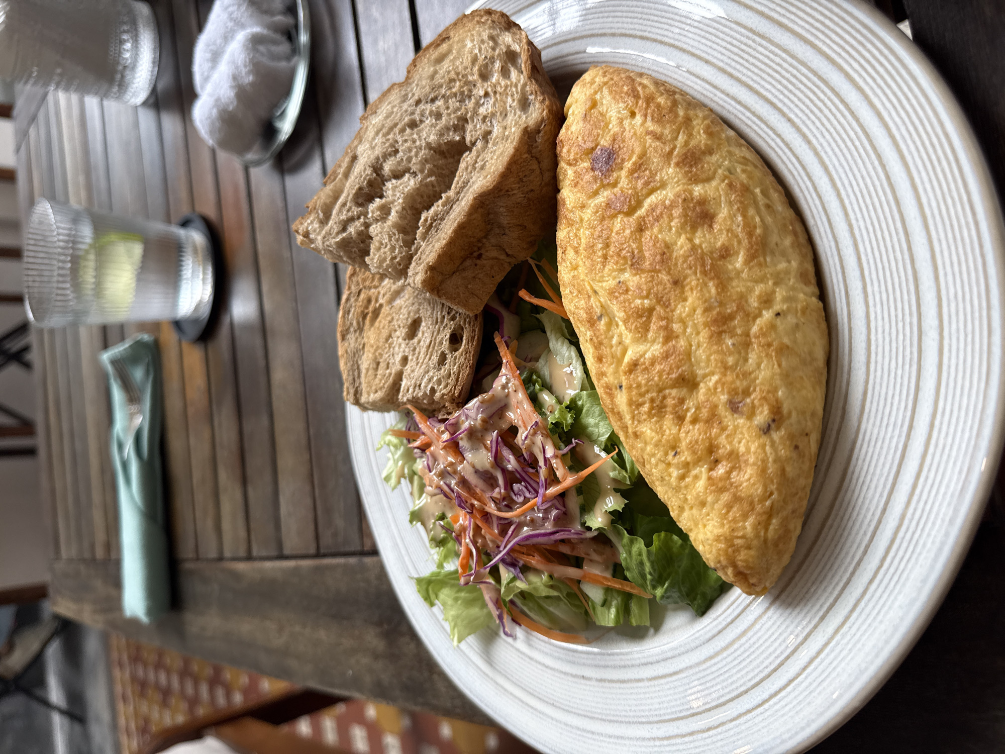 A plate of food sits on a dark wooden table. The plate contains an omelet, a salad with shredded carrots and purple cabbage, and two slices of toast. To the side of the plate is a glass of water with a lime wedge, a small folded cloth, and a small bowl of cotton balls. The image appears to have been taken in a restaurant or cafe in Đà Nẵng, Vietnam.