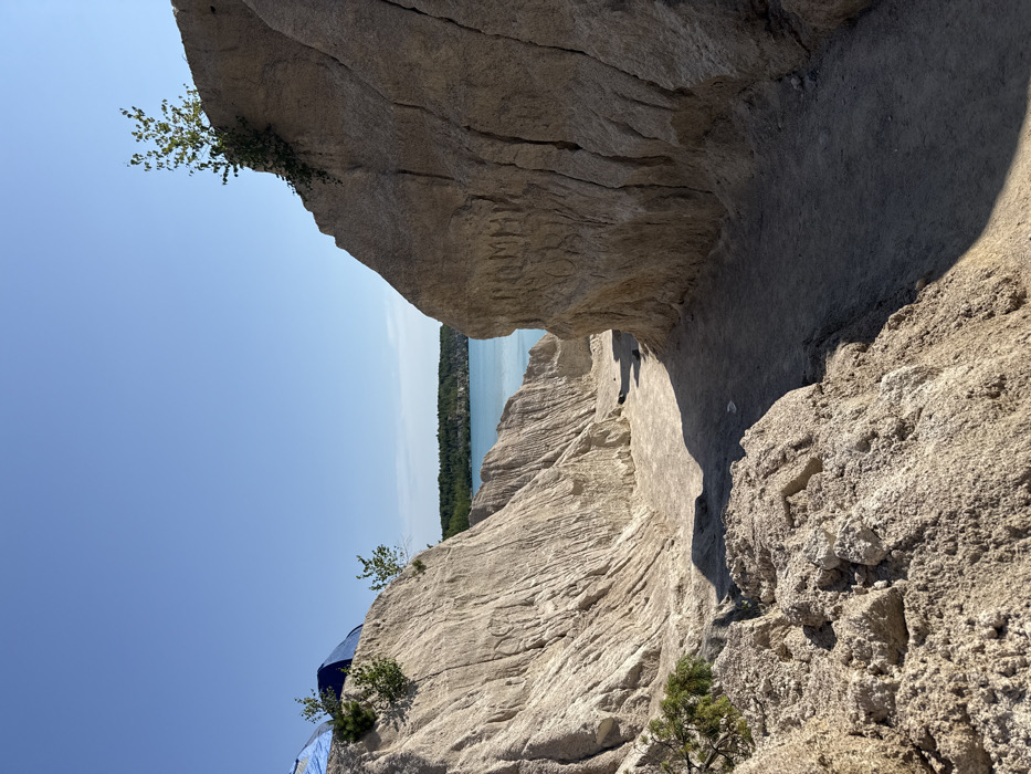The image presents a sunlit outdoor scene within the former chalk or sand quarry near Druzhba, Ukraine, characterized by a unique landscape. In the foreground, a narrow, unpaved dirt path recedes between two prominent, steep cliffs composed of light-colored, heavily eroded material, exhibiting vertical striations and varied textures. The path's surface shows areas of shadow from the elevated cliff walls. On the upper left side, situated atop a lighter-colored ridge, two blue camping tents are visible, partially obscured by small, green vegetation. Additional green plant growth, including small trees or bushes, emerges from the surfaces of both the left and right cliff faces. Notably, the lower section of the right cliff displays carved Cyrillic lettering, which appears to spell "ПОМОГ" (meaning "helped" or "assisted"). Beyond the immediate geological formations, a large body of vibrant turquoise-blue water extends into the mid-ground, backed by a dense, dark green line of deciduous forest. The sky above is clear and uniformly blue, with no visible clouds. The scene is illuminated by direct sunlight, casting distinct shadows on the ground and cliff surfaces. No individuals are directly visible.