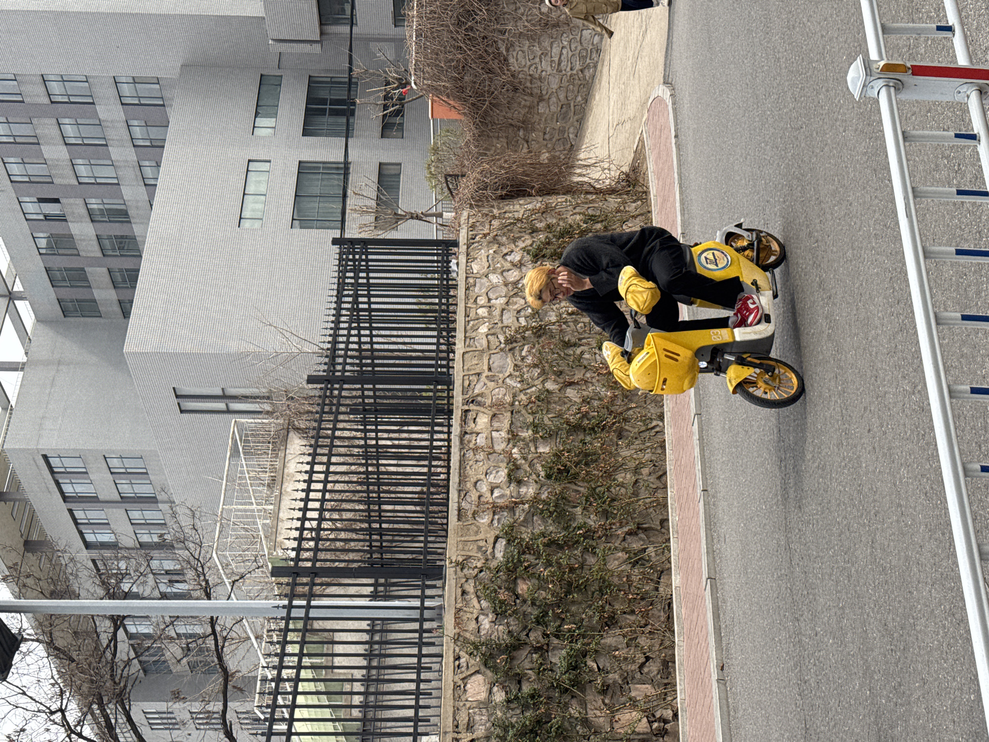 A person is on a yellow scooter next to a road. Adjacent to the road is a stone wall. To the left of the wall is a fence and a building. The person appears to be looking at something on the scooter. In the background is a building, and on the far right is a white metal barrier. The location is Zhongtun, China.