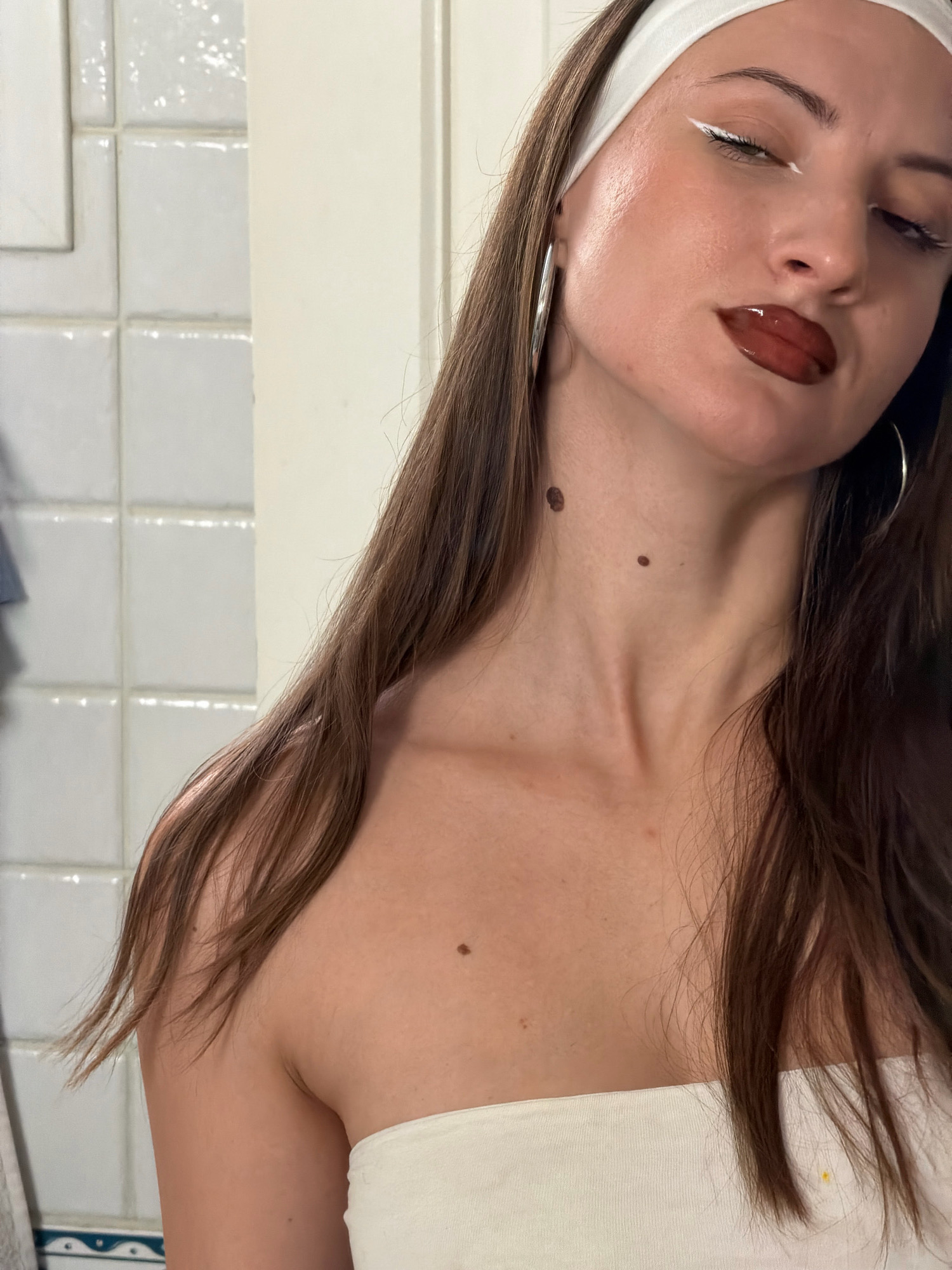 The image is a close-up portrait of a young woman, seen from the chest up, against a tiled bathroom wall. She has long, dark brown hair and is wearing a white headband and a white strapless top. Her makeup includes bold white eyeliner, dark lipstick, and subtle blush. She has several moles visible on her neck and chest. The lighting suggests an indoor setting, likely a bathroom, with bright lights reflecting off the tiles and her skin. There are no discernible landmarks or elements that would place this image specifically in Sharm El Sheikh, Egypt.