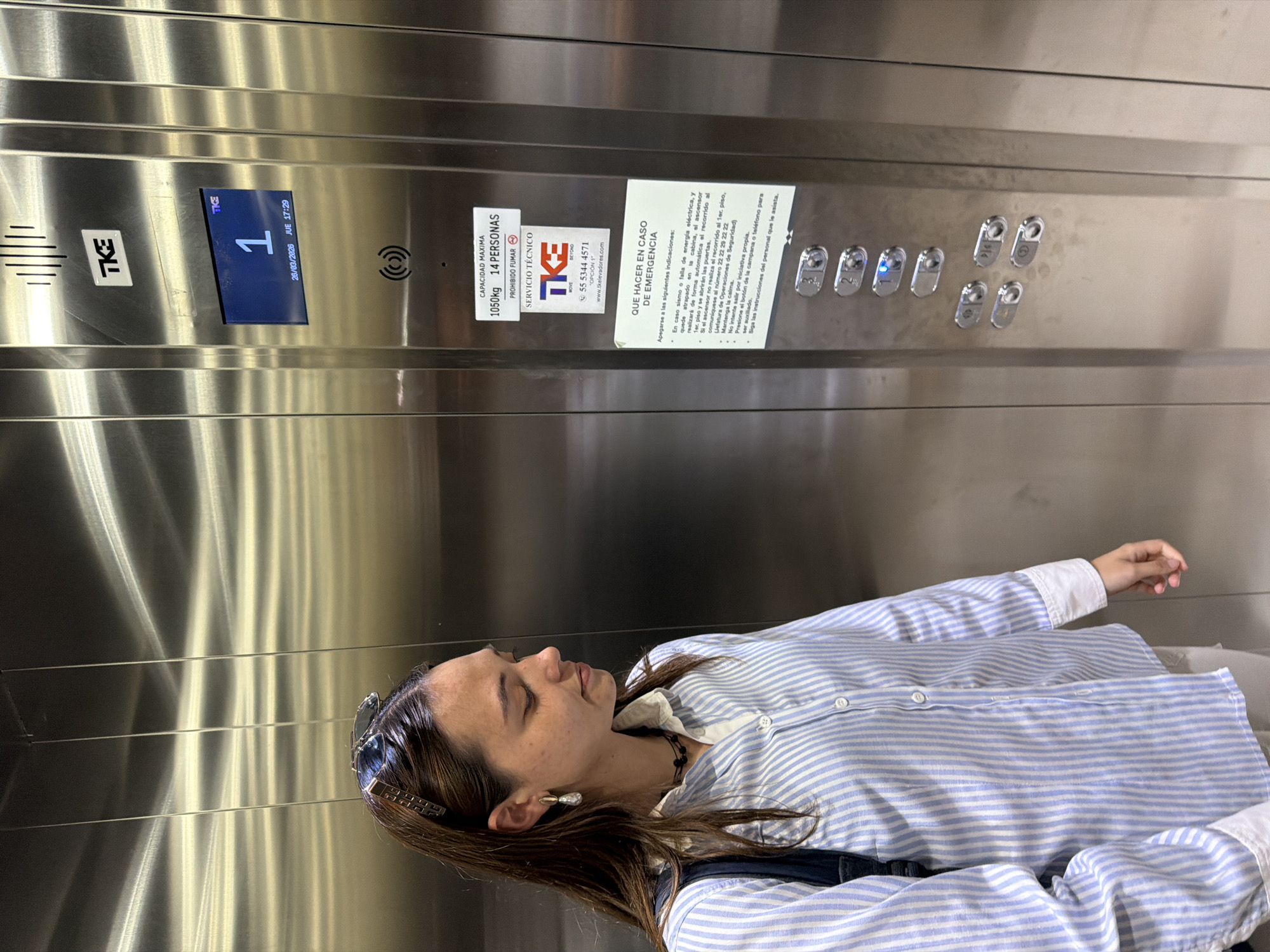 The image depicts the interior of a TKE-branded elevator with brushed stainless steel walls, located in San Andrés Cholula, Mexico. The digital display indicates the elevator is on floor "1" and shows the date as March 26, 2026, at 17:29 (5:29 PM).
A single female individual occupies the left side of the frame. She has long, dark brown hair, with sunglasses and a gold-colored hair clip resting on top of her head. She wears teardrop-shaped earrings, a dark beaded necklace, and a light blue and white vertically striped long-sleeved button-down shirt with white cuffs. A dark-colored backpack strap is visible on her left shoulder. Her head is slightly bowed, and she is looking downwards and to her right (viewer's left), with a neutral expression. Her right hand hangs loosely at her side.
The elevator control panel, located on the right, features the digital display at the top, followed by a TKE logo. Below this are white stickers providing information in Spanish: "CAPACIDAD MAXIMA 1050kg 14 PERSONAS" (Maximum Capacity 1050kg, 14 Persons) and "PROHIBIDO FUMAR" (No Smoking). A "SERVICIO TÉCNICO" (Technical Service) sticker displays the TKE logo, a phone number (55 5344 4571), and a website. Further down, a sign titled "QUE HACER EN CASO DE EMERGENCIA" (What to do in case of emergency) lists instructions in Spanish. A vertical array of chrome-finished buttons is visible below the signs, with the "1" button illuminated in blue, indicating the current floor or a selected destination. The metallic surfaces of the elevator show horizontal light reflections.