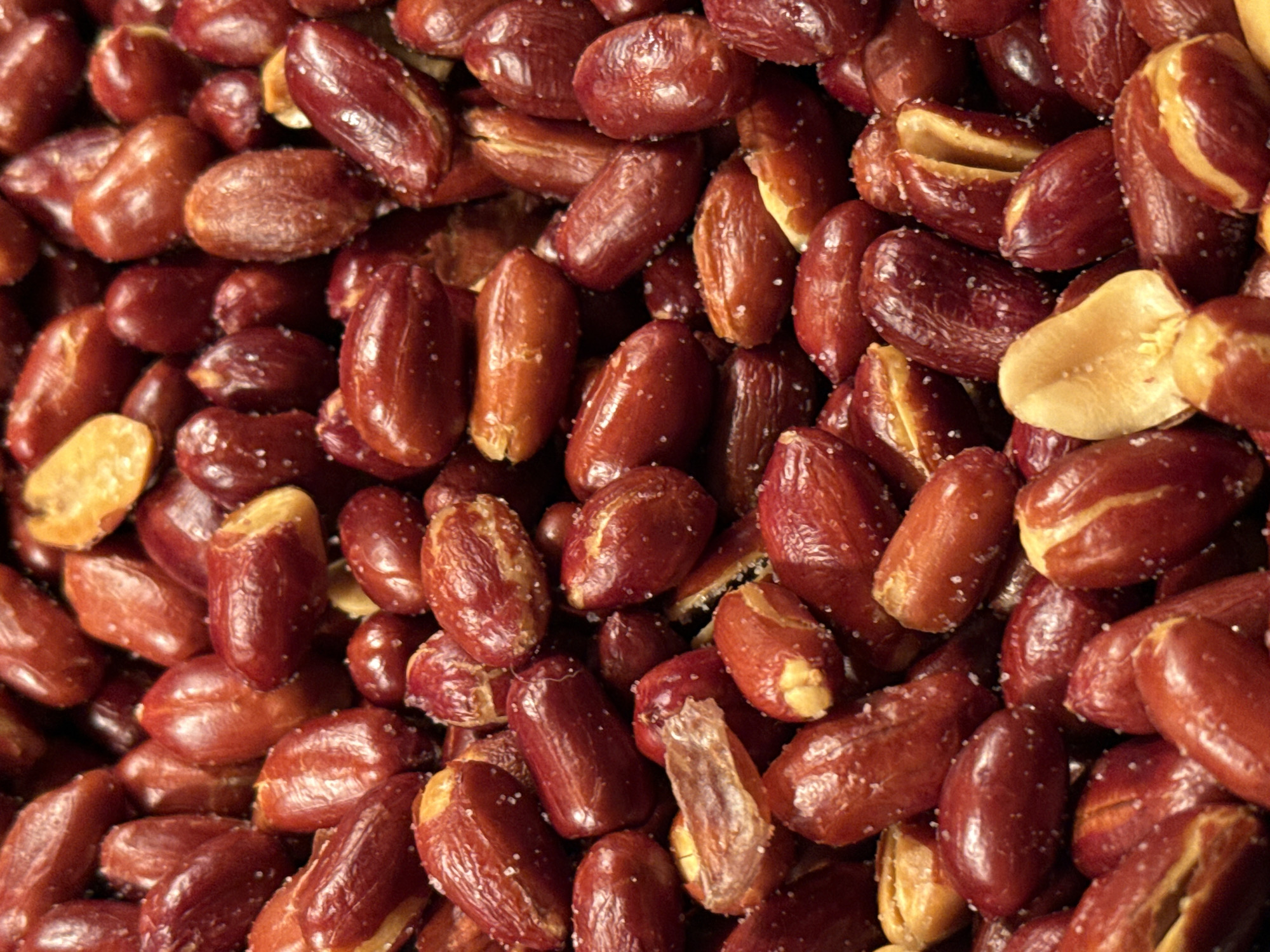 The image is a close-up, overhead shot of a pile of roasted peanuts. The peanuts are predominantly reddish-brown with some lighter tan patches where the skins have split open. They appear to be salted, with small white flecks of salt visible on their surfaces. The lighting is somewhat dim, creating a rich, warm tone and highlighting the textures of the peanuts. The focus is sharp, and the background is blurred, emphasizing the abundance of peanuts in the foreground. There are no people, actions, or environmental elements identifiable within the image. The scene is entirely composed of roasted peanuts.