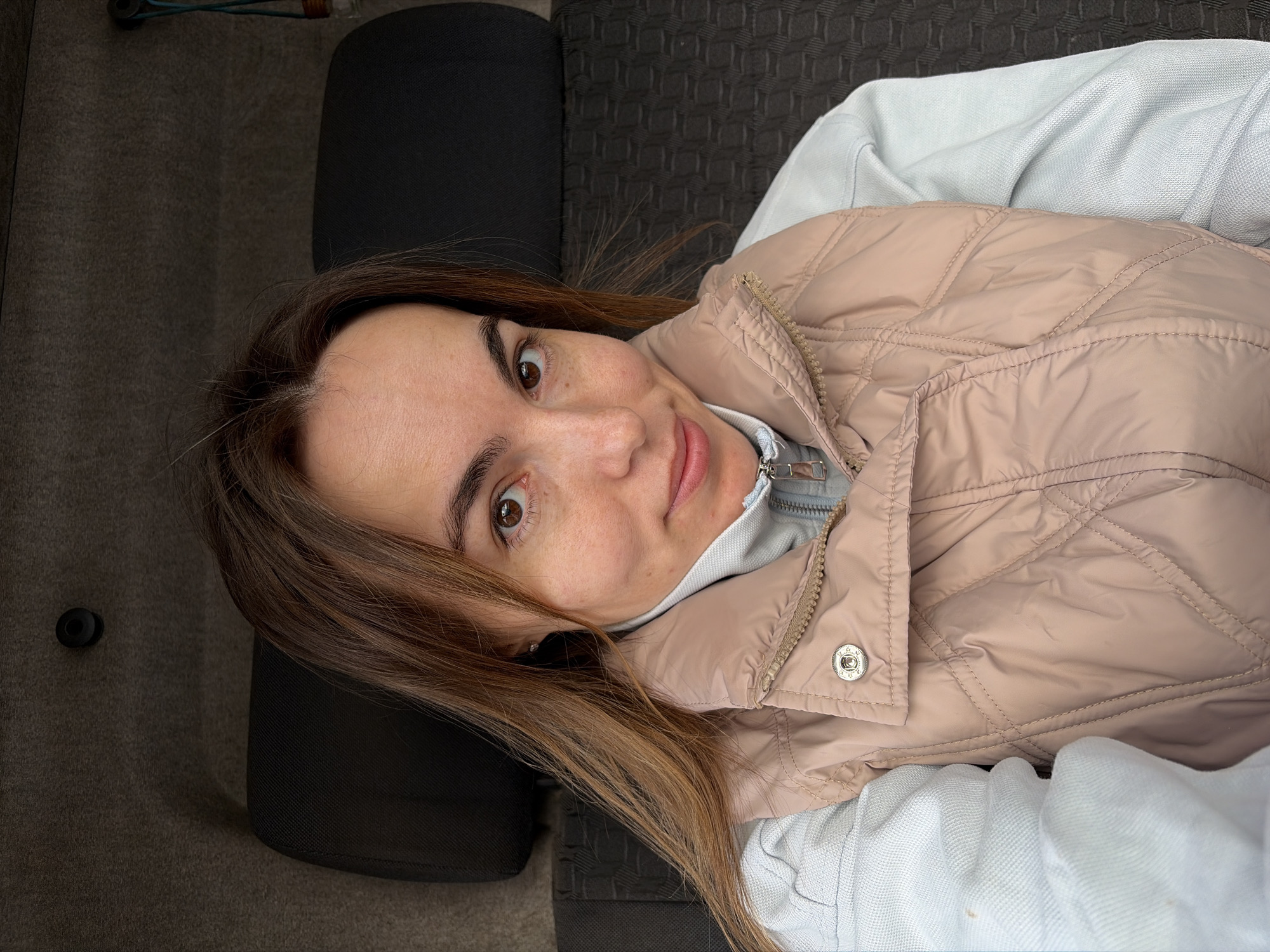 A woman with brown hair and brown eyes is lying down, wearing a light blue hooded sweatshirt and a light pink quilted vest. She is looking at the camera with a slight smile. She is lying on what appears to be a grey upholstered surface next to a black upholstered object. The location is not discernible from the image.