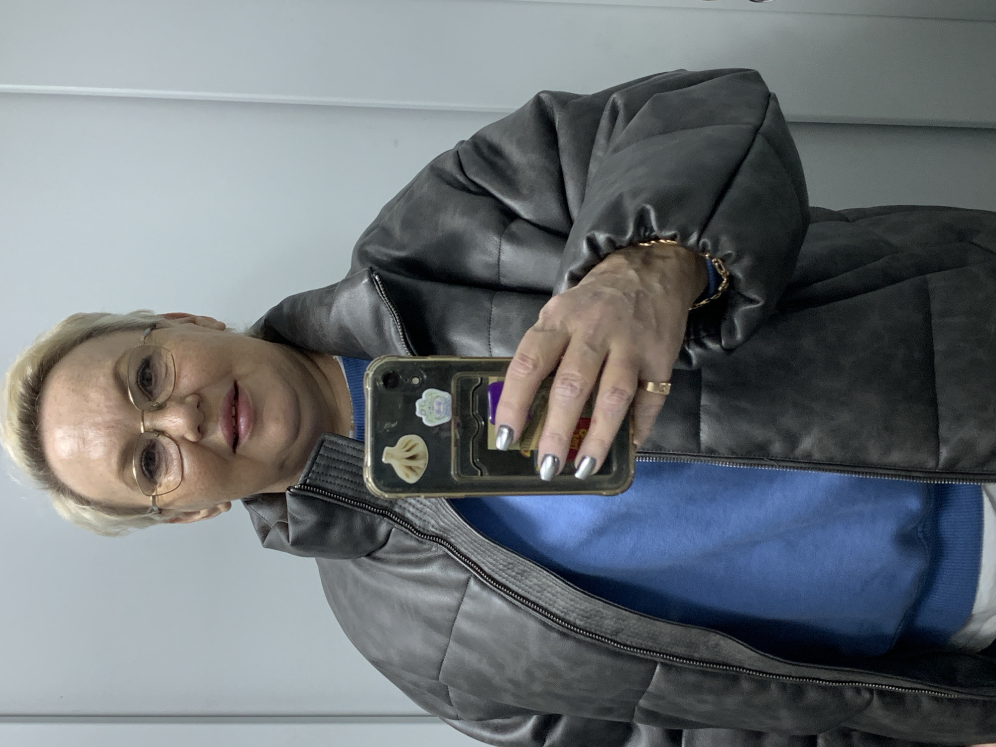 An individual is captured in a mirror selfie taken indoors in Chaikovsky, Russia. The subject, a woman with short blonde hair and gold-rimmed glasses, is seen from the chest up, with a slight smile on her face. She wears a dark gray or black padded puffer jacket, which is partially unzipped to reveal a bright blue long-sleeved top underneath, likely a sweater.
She holds a smartphone in her right hand, featuring a clear case adorned with several stickers, including one shaped like a dumpling or khinkali and another small cartoon character. A purple card, possibly a bank card with "Сбербанк" faintly visible, is tucked into the back of the phone case. Her fingernails are painted silver, and a gold ring is worn on her right ring finger. A thin gold chain bracelet is visible on her left wrist, peeking from under the jacket sleeve. The background is a plain, light-colored wall, indicating an uncluttered indoor setting. The image is oriented sideways, suggesting the phone was held horizontally or the original capture was rotated.