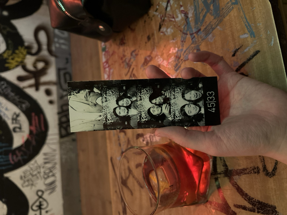 A hand holds a photo booth strip featuring a couple. The strip is divided into multiple black and white images. The number "45|33" is printed on the far right. The images are slightly obscured by a glass of reddish liquid. The setting appears to be a bar, with a graffiti-covered wall and painted wooden surface visible. A black purse is positioned in the upper left corner.