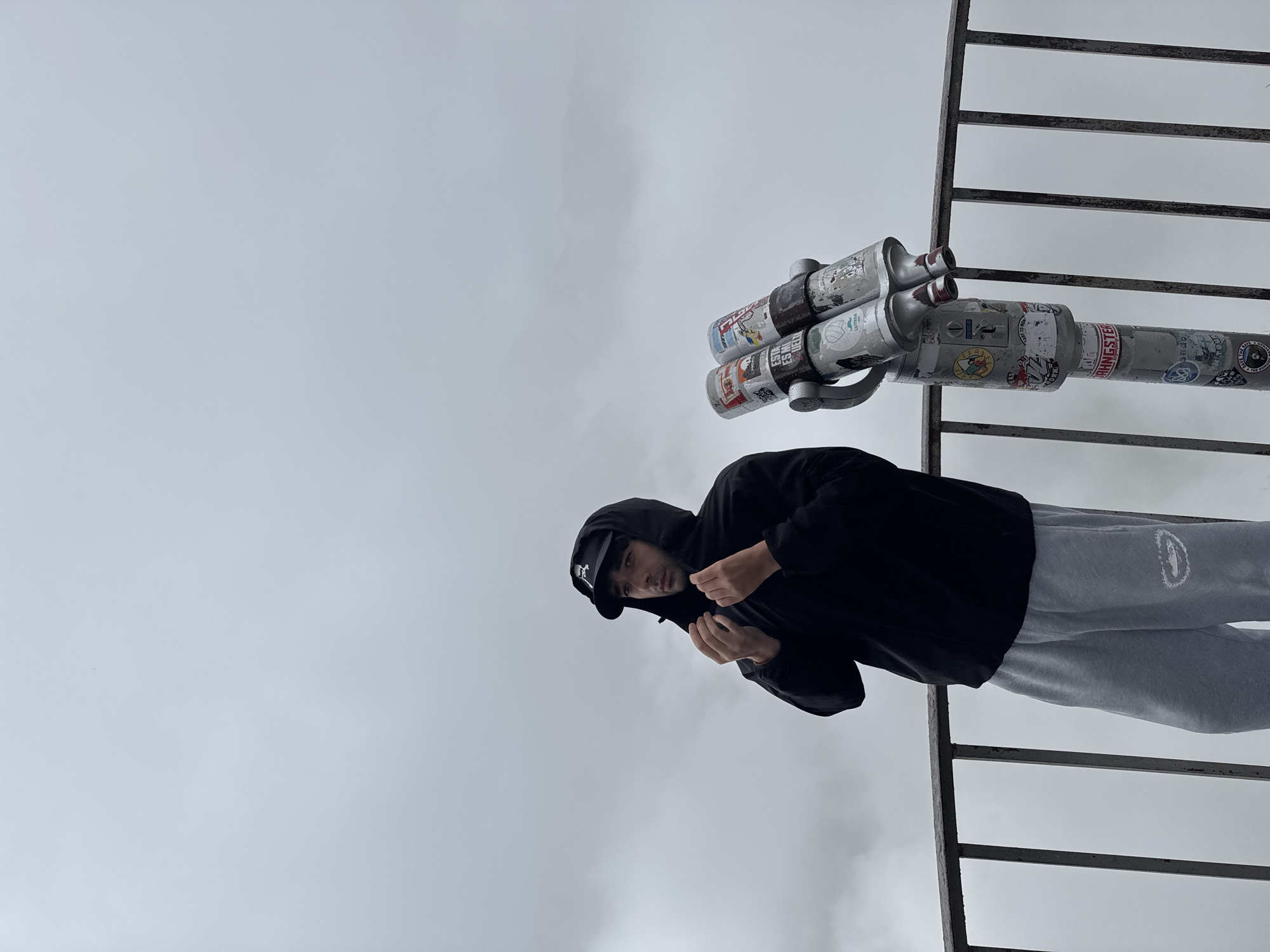 A man stands on an observation deck, leaning on a railing. He is wearing a black hooded jacket and light gray sweatpants. The hood of his jacket is pulled up, and he is adjusting the drawstring. To his right is a pair of coin-operated binoculars covered in stickers. The man is looking to his left, away from the camera. The background is a solid gray sky, indicating an overcast or foggy day. The location is the Gudauri viewpoint in Gudauri, Georgia, famous for its stunning mountain vistas, which are currently obscured by the weather.